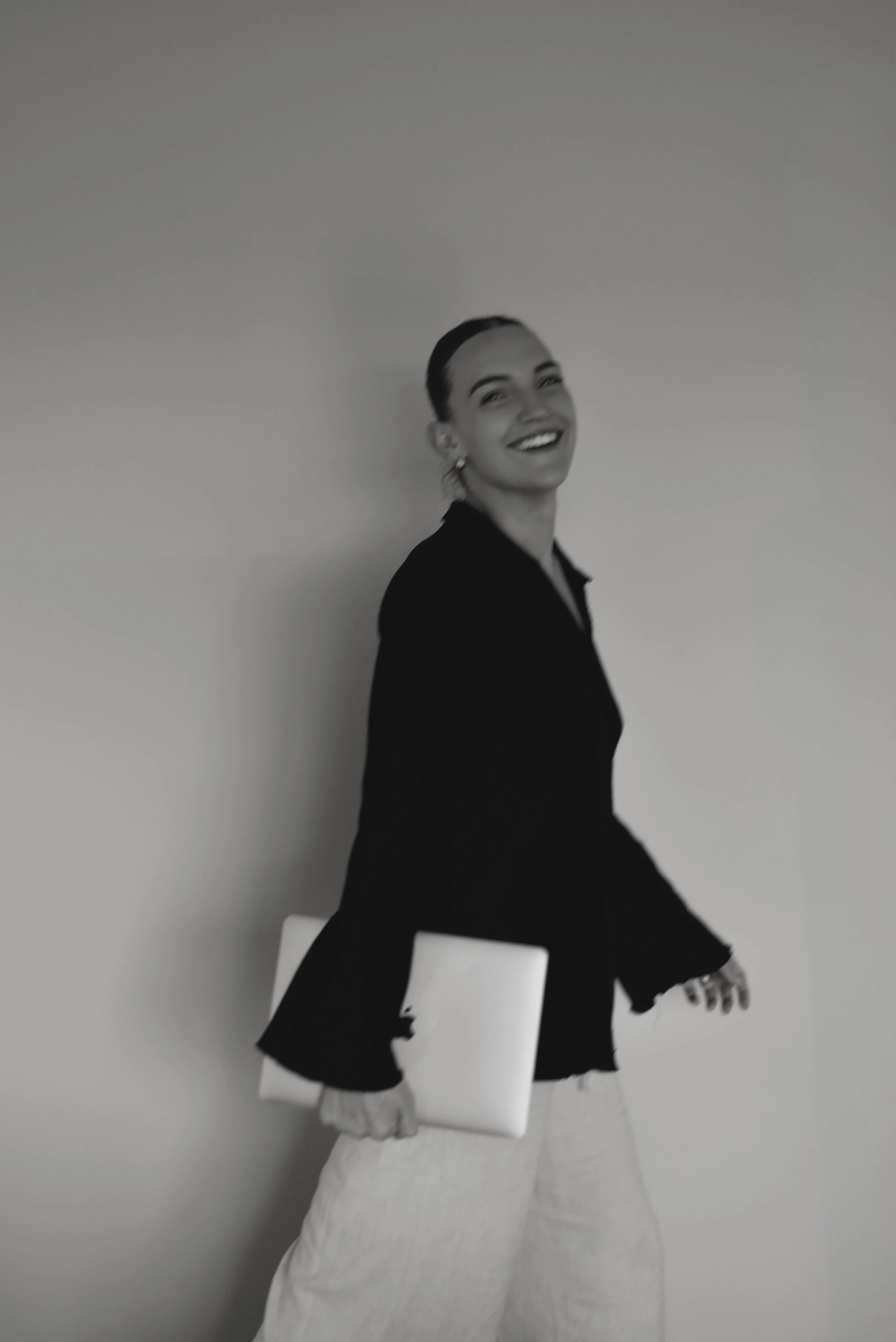 A woman smiling, wearing a dark blazer and light-colored pants, holding a closed laptop in her right hand, standing against a plain wall.