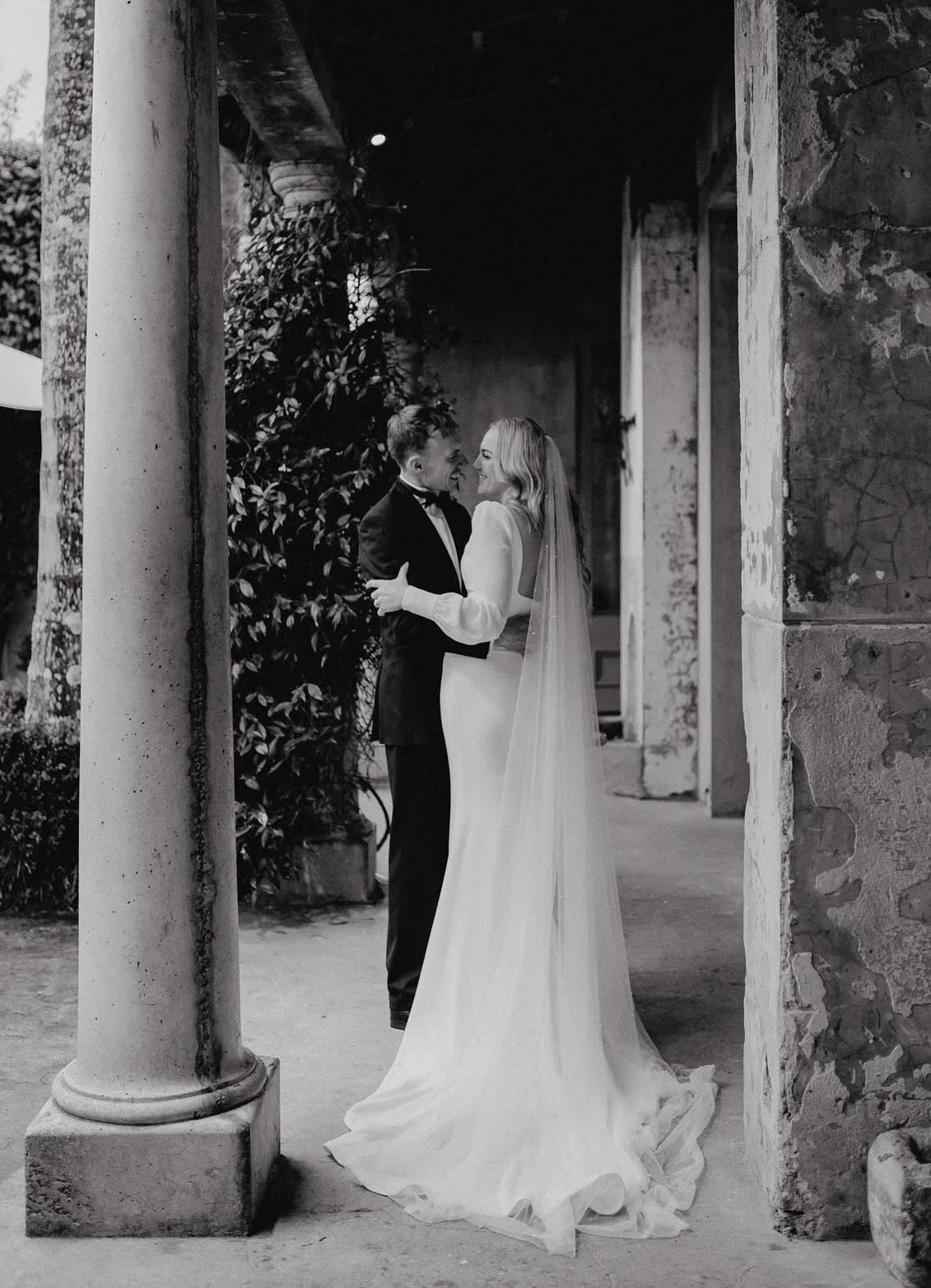 A black and white photo of a bride and groom smiling and looking at each other, standing close under a stone archway with columns and greenery in the background.