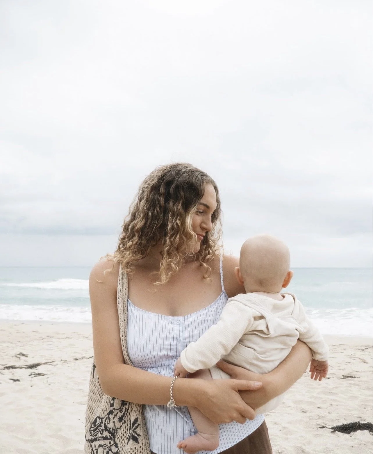 A woman holding a baby at the beach with the ocean and cloudy sky in the background.