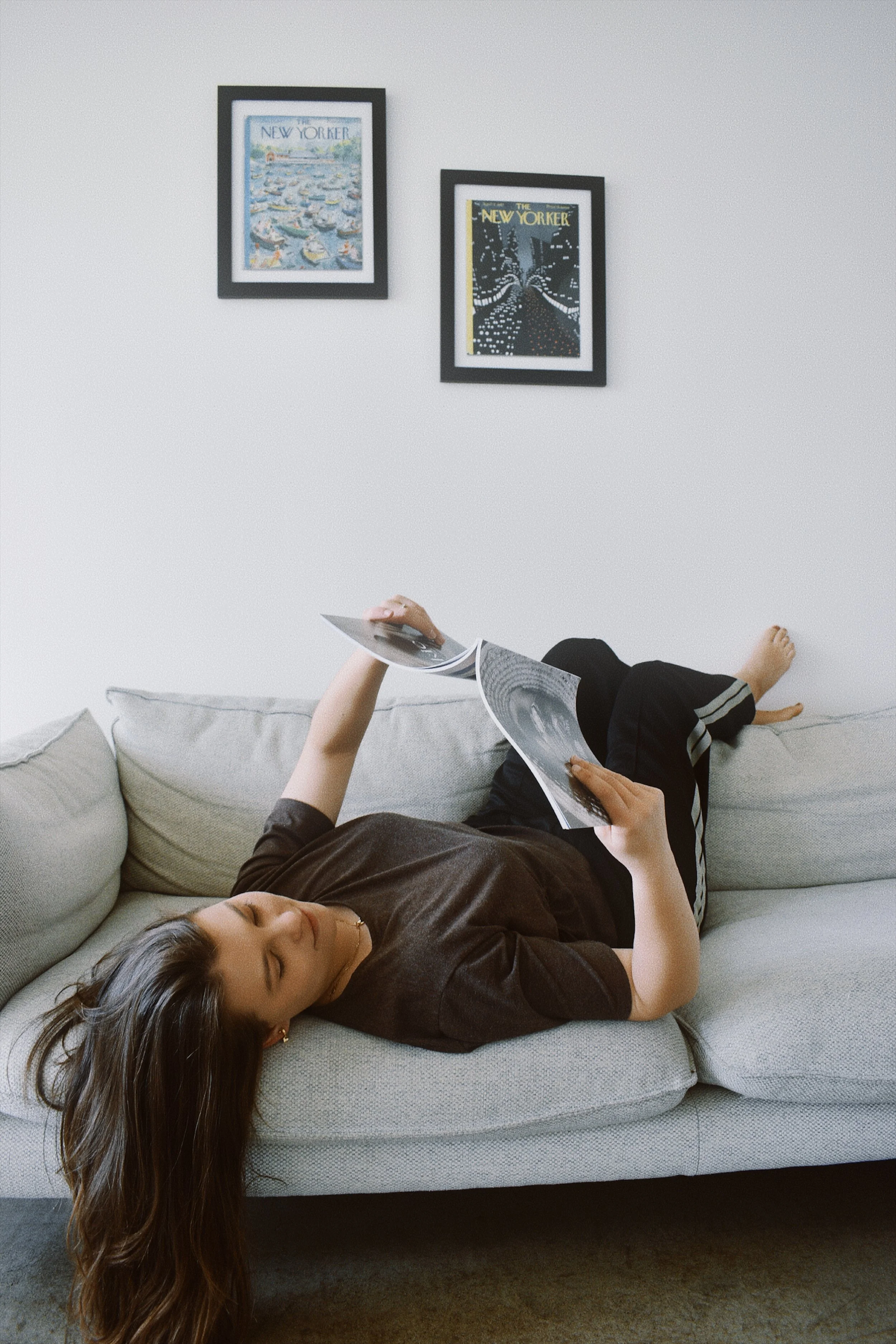 A woman is lying on a light-colored sofa, reading a magazine with black and white images. She is wearing a dark t-shirt and black shorts with white stripes. There are two framed art prints on the white wall above her, one depicting boats and the other a cityscape.