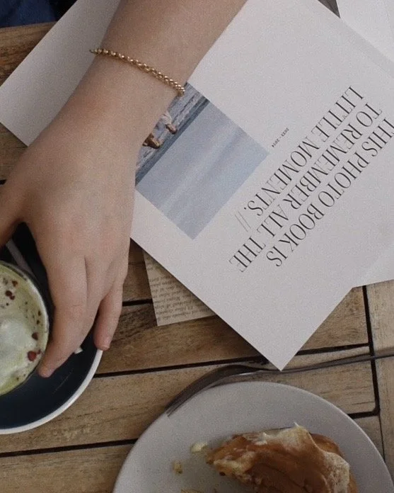 A person's hand with a gold bracelet reaches for a cup of coffee, placed on a wooden table with a newspaper, a plate with a piece of pastry, and a small stack of papers.
