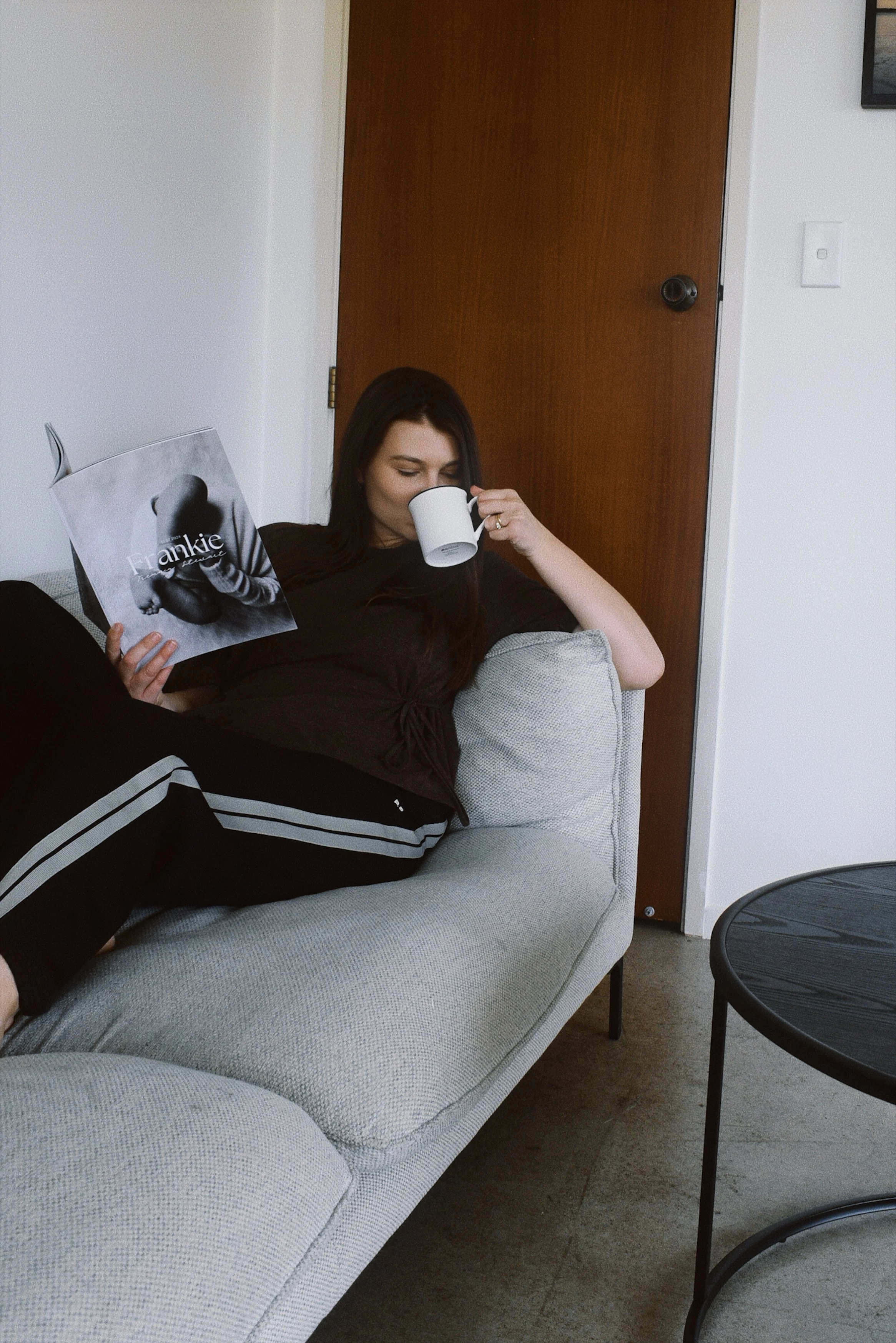 A woman with long dark hair is relaxing on a light gray sofa, reading a magazine titled 'Frankie' and drinking from a white mug in a living room with a closed wooden door behind her.