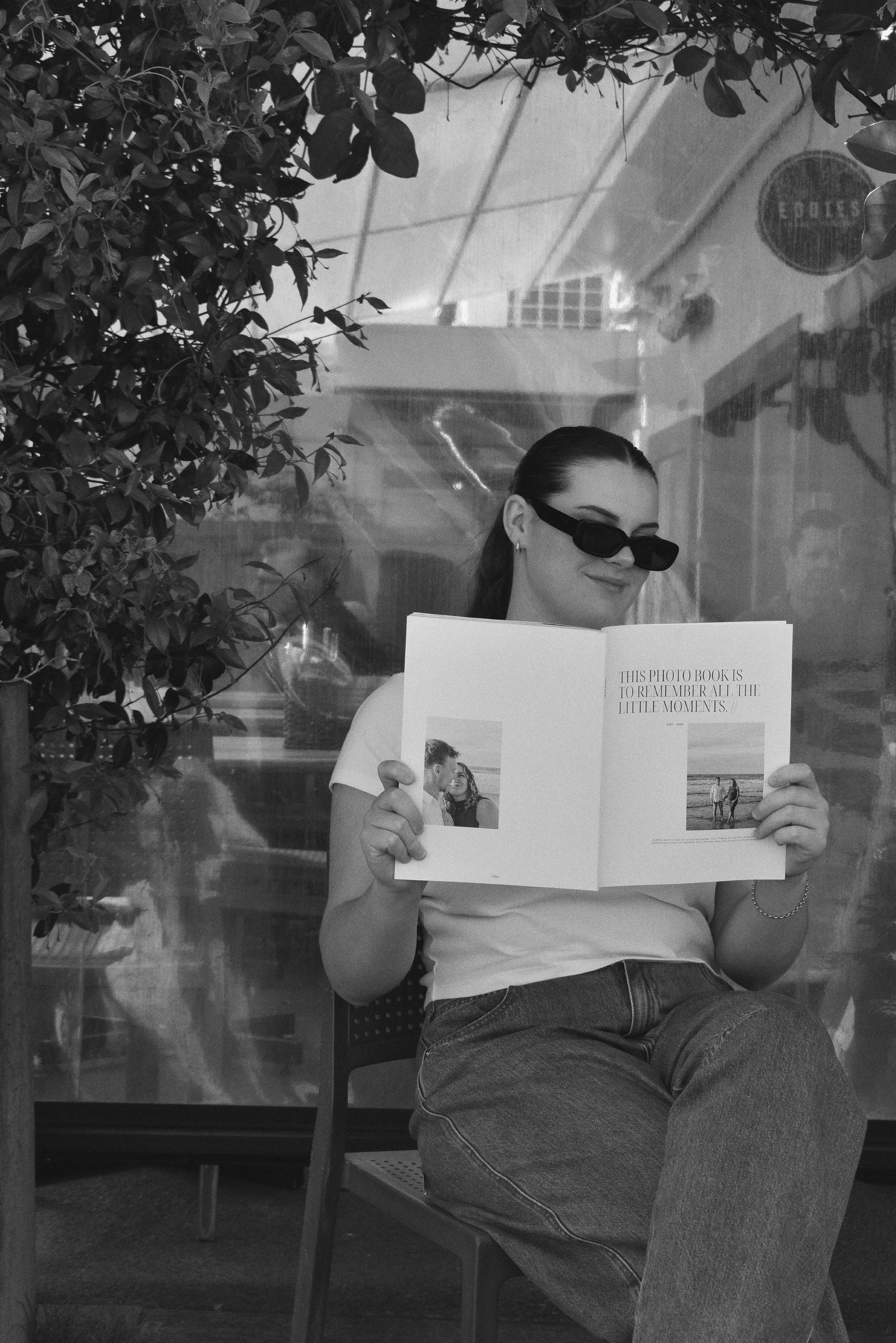 A woman with dark hair, sunglasses, and jewelry sits on a chair, reading a photo book, with a leafy plant to her left and an indoor setting in the background.