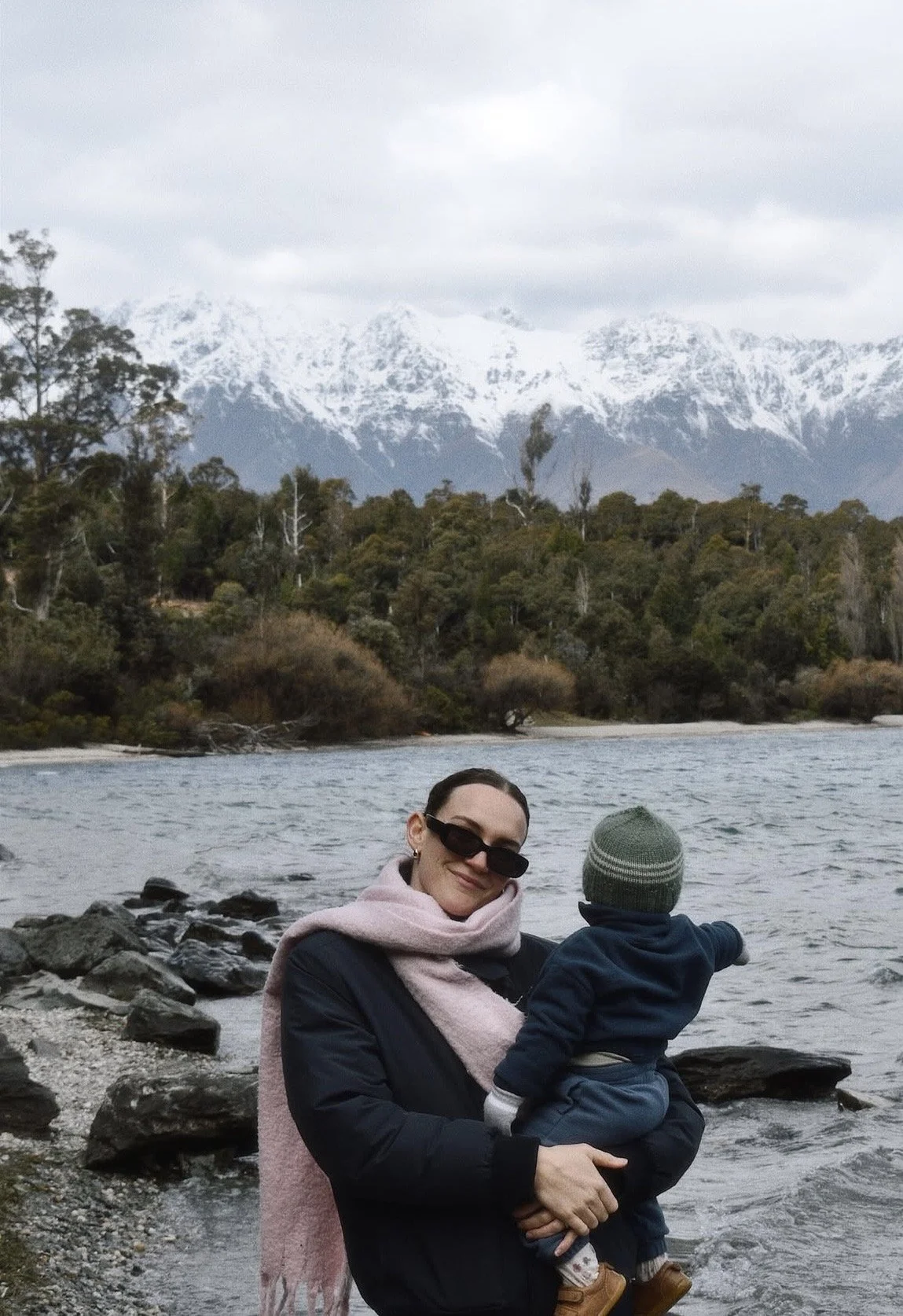 A woman wearing sunglasses, a pink scarf, and a black jacket holds a young child in front of a river with snow-capped mountains and trees in the background.