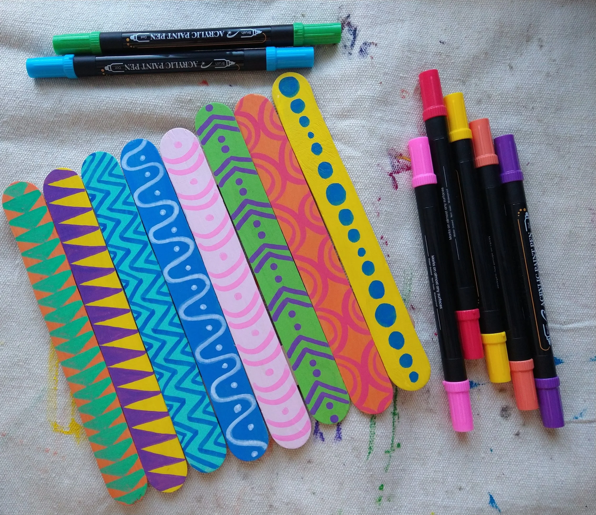 Une rangée de marqueurs de différents couleurs avec des capuchons, accompagnée de plusieurs marqueurs à pointe large, tous posés sur un tissu blanc avec des traces de peinture