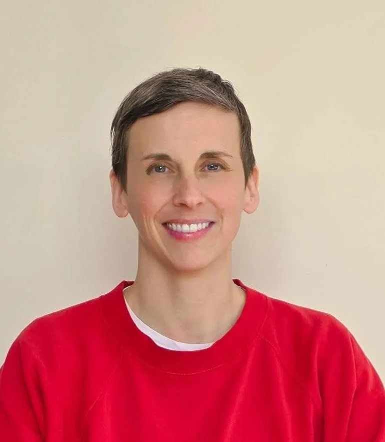 Une femme souriante aux cheveux courts, portant un sweat rouge, devant un mur beige clair.