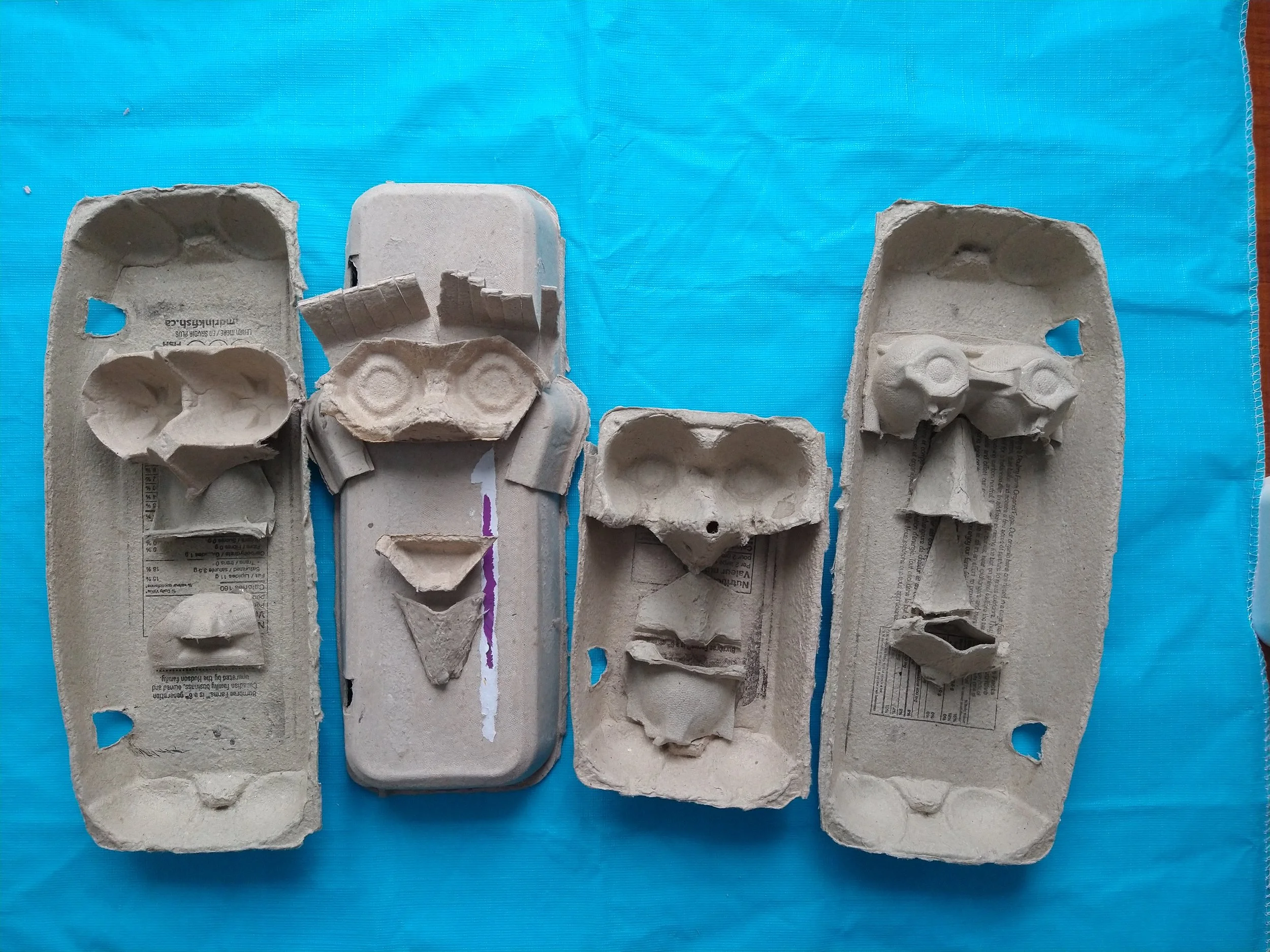 Quatre plateaux en carton d'œufs, certains contenant des formes en œuf cassé ou en carton d'œufs, disposés sur une surface en plastique bleu.