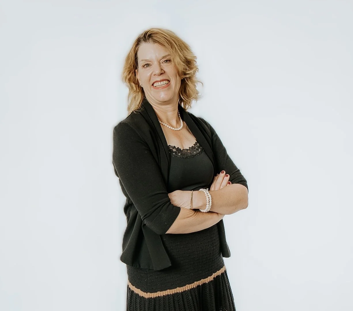 Julie Perron — a woman with wavy blonde hair smiling with arms crossed, wearing a black blazer, black dress with lace details, pearl necklace, and bracelets, standing against a plain white background.