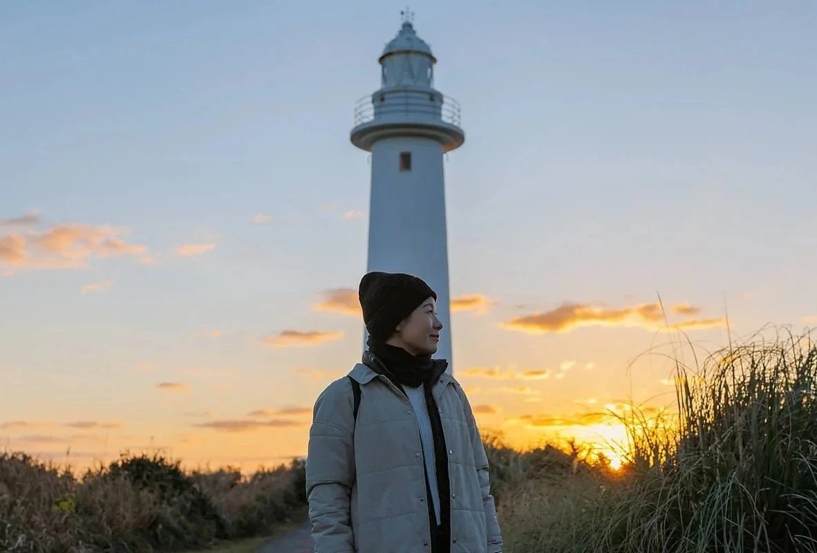 Smiling woman standing in front of a lighthouse during sunrise, symbolizing the insightful, reliable actuarial support provided by Ordinance.
