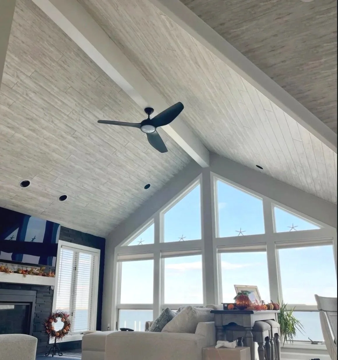 Living room with a vaulted ceiling with shiplap, ceiling fan, large windows, and a fireplace with a wreath above it.