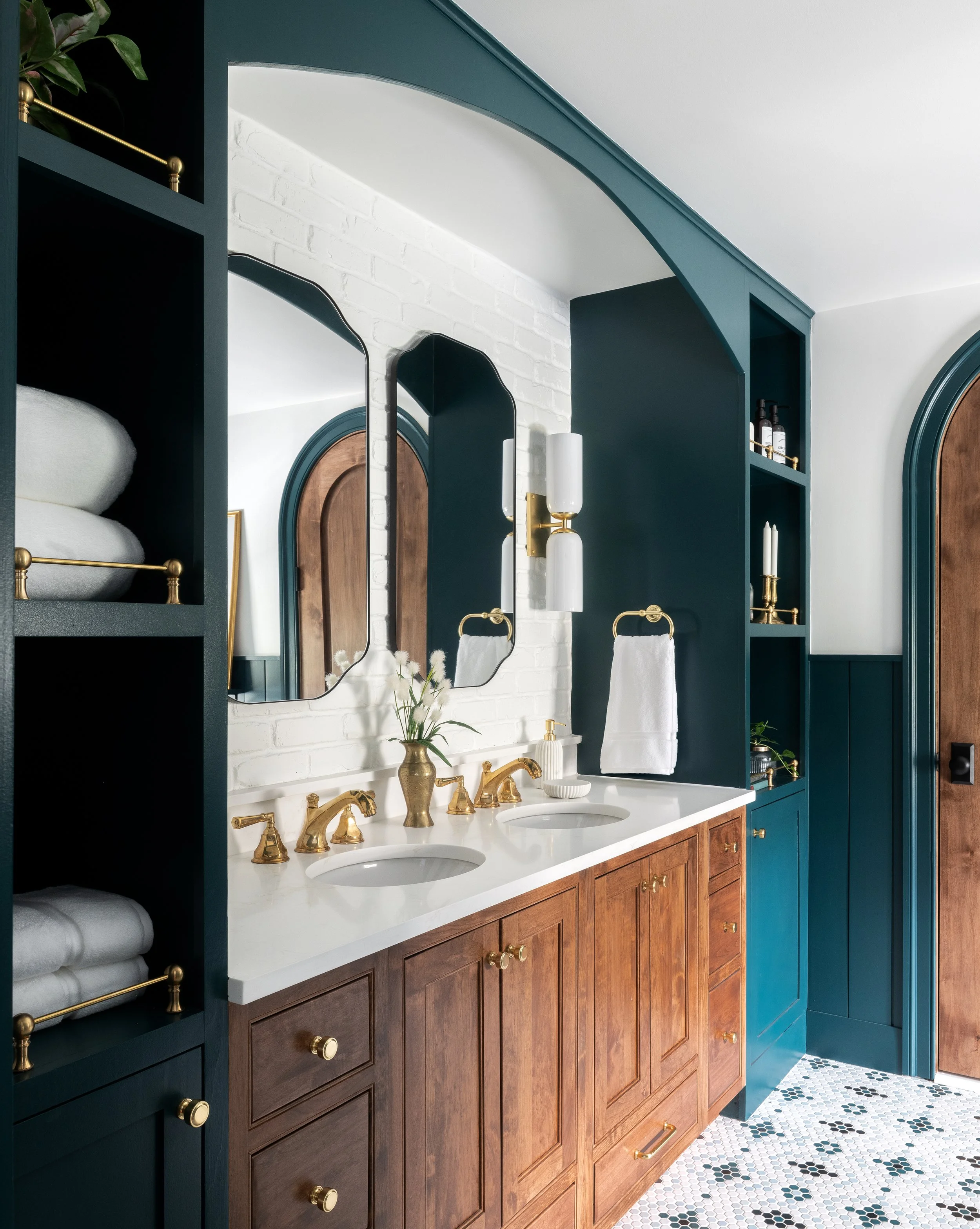 Modern bathroom with a double sink vanity, wooden cabinets, gold fixtures, and vibrant teal walls. Features decorative mirrors, wall-mounted lights, shelving with towels and accessories, and a stage-patterned tile floor.