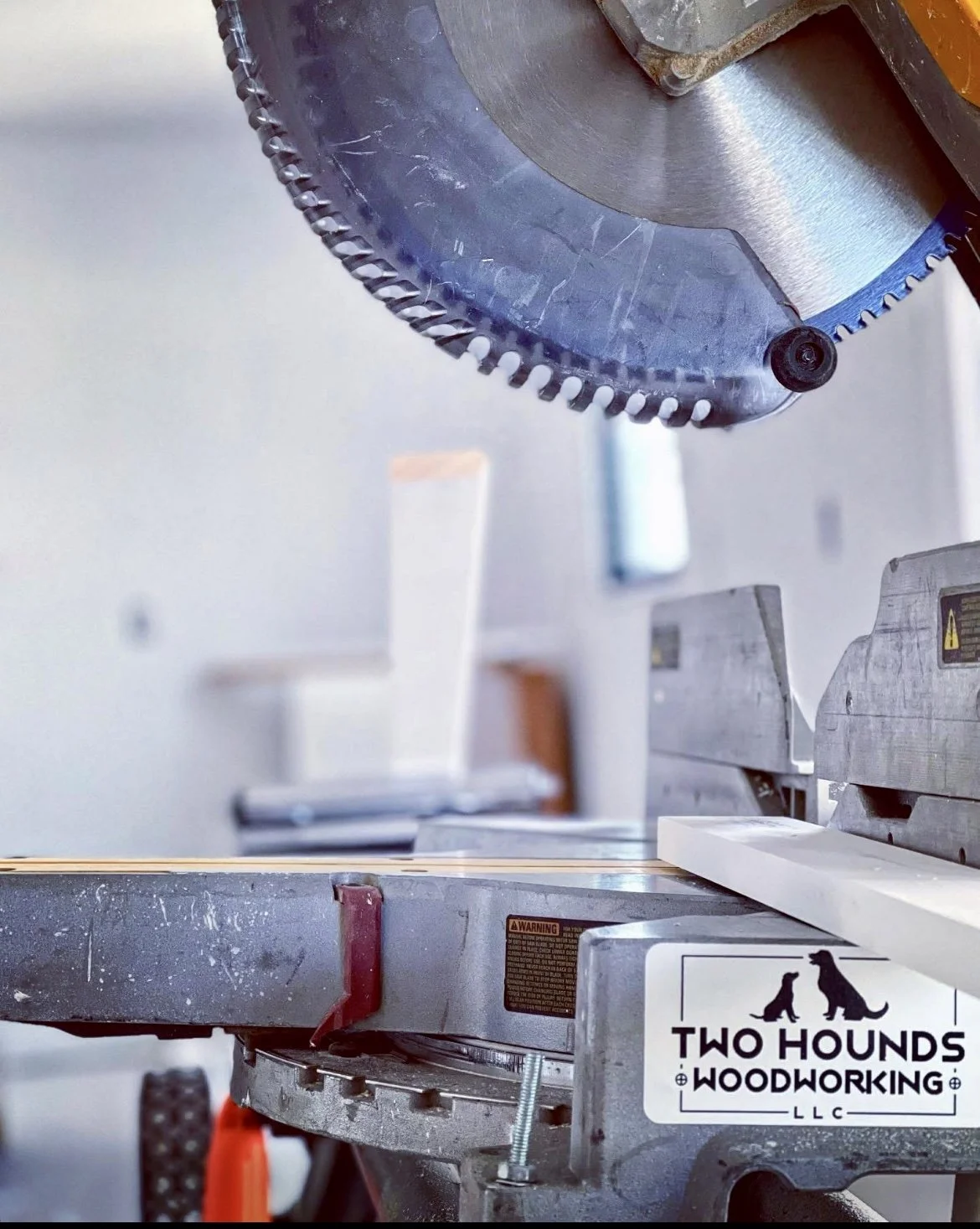Close-up of a circular saw blade on a woodworking machine in a workshop. The saw blade has sharp teeth and is mounted on an orange and gray machine. There is a sticker on the machine that reads 'Two Hounds Woodworking LLC' with silhouettes of two dog
