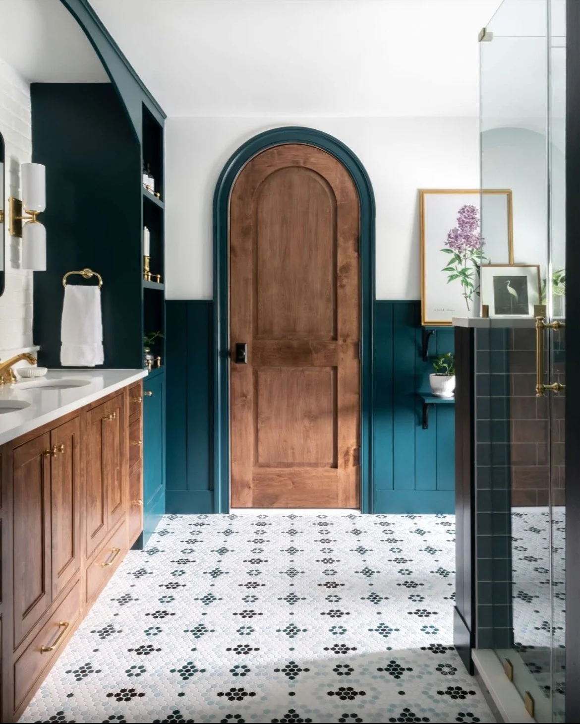 Modern bathroom with custom wooden cabinets, teal wainscoting, and a custom wooden trimmed arched door. Features include framed art with flowers and a bird, a plant, and a glass shower door.