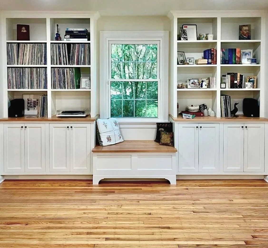 White built-in bookshelf with vinyl records, books, framed photos, and decorative items, centered around a window with view of trees, on a hardwood floor.