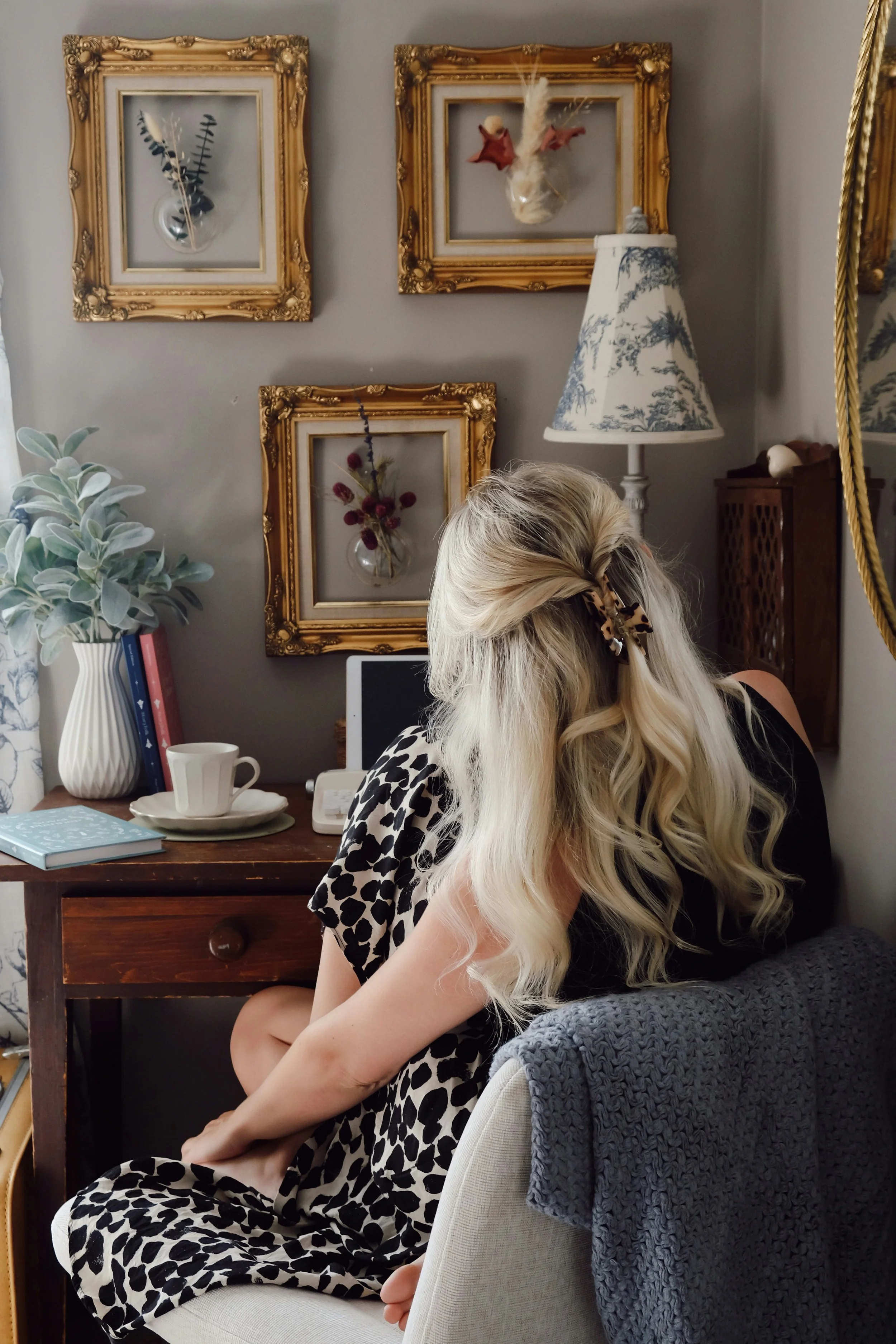 Une femme blonde coiffée d'un chouchou à motif animal, assise dans un salon vintage, face à un ordinateur portable, entourée de décorations rétro, dont une lampe, un vase avec des feuilles, des livres et des tableaux encadrés ornant le mur.