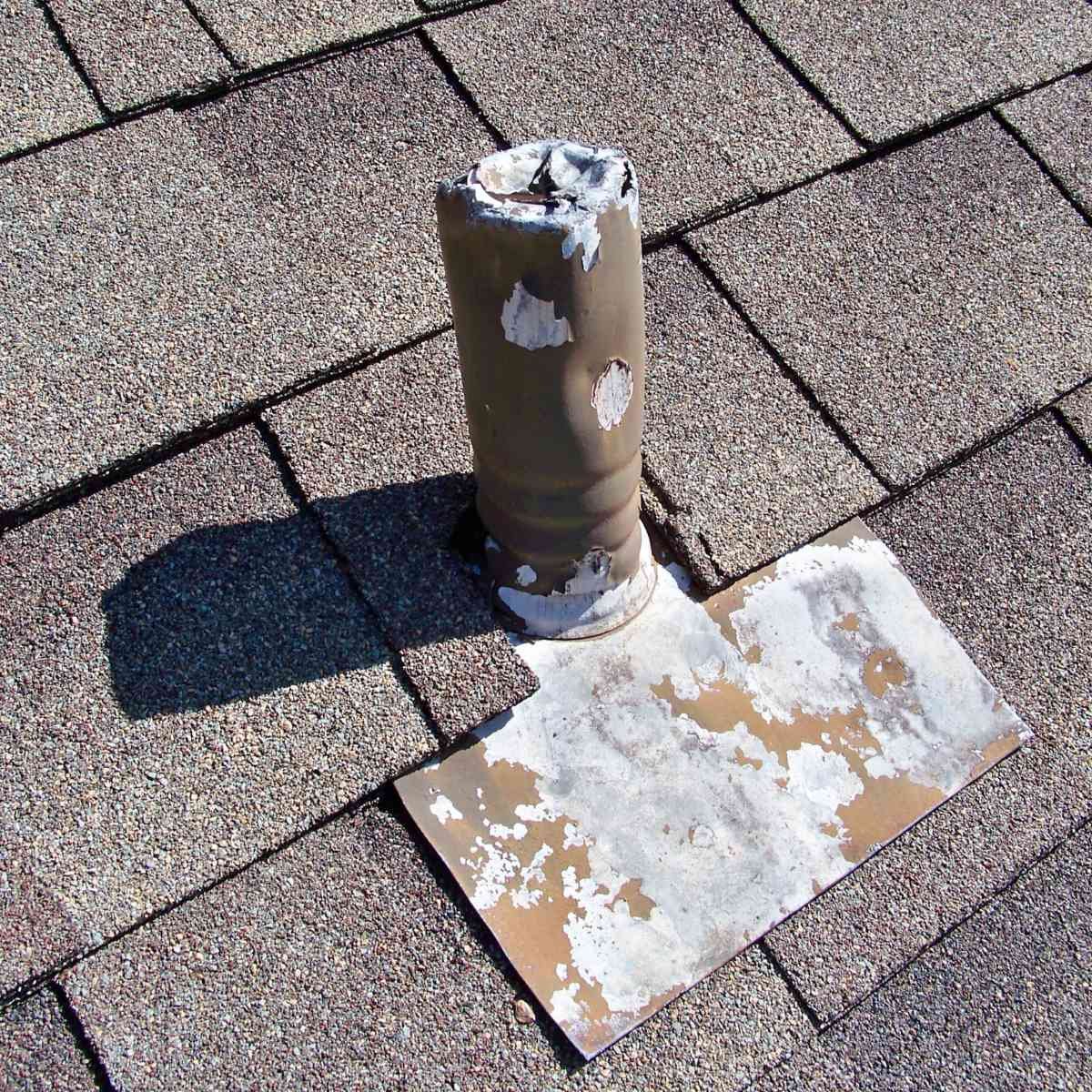 Old, weathered metal pipe protruding from a roof, with peeling white paint, casting a shadow on the shingles. A square patch of peeled paint or primer is nearby.