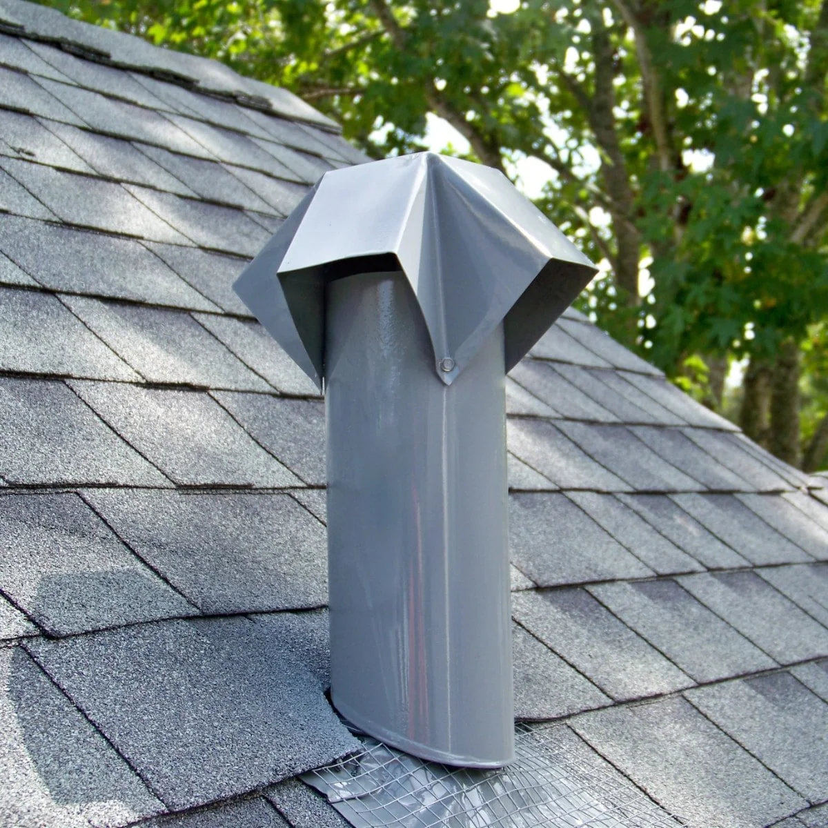 Metal chimney cap installed on a shingled roof with trees in the background.