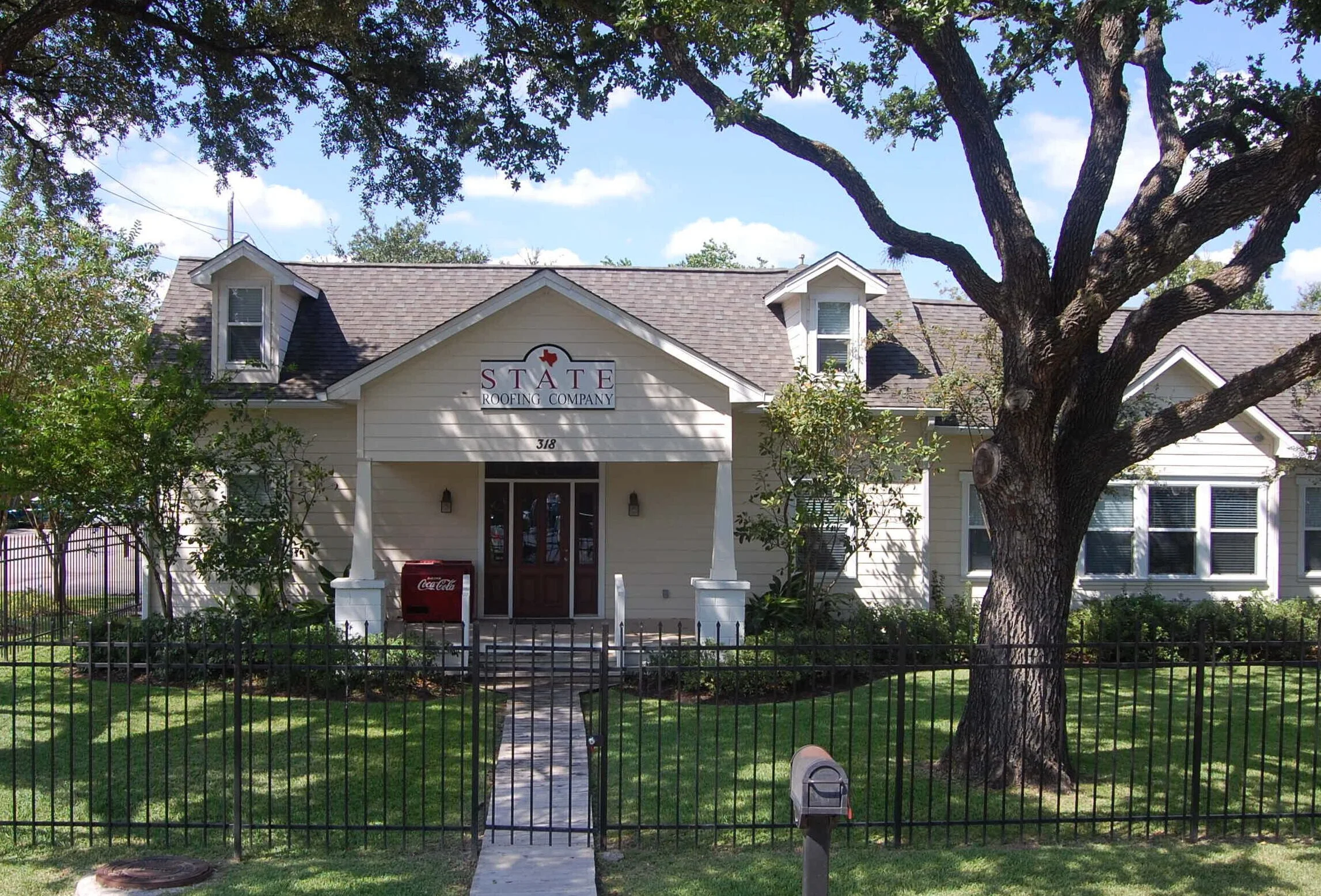 A small, cream-colored building with a gabled roof and a porch, surrounded by a black metal fence. There is a large tree in front of the building. The building has a sign that reads 'STATE ROOFING COMPANY' with a Texas-shaped logo. The address number 318 is on the front of the building.