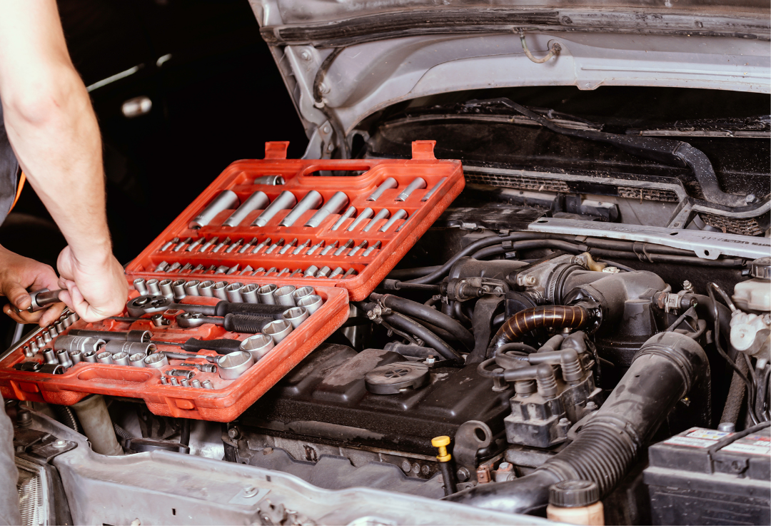 A person working on a car engine with a red toolbox containing various socket tools, with the car's hood open.