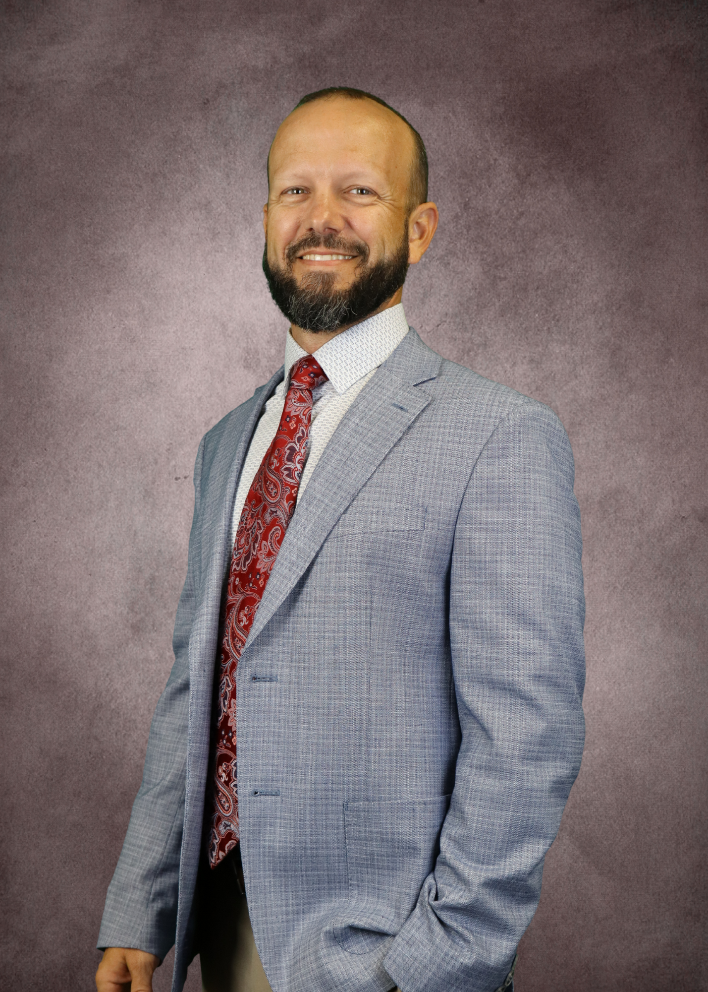 A professional man with a beard, smiling, wearing a light gray suit, white shirt, and a red paisley tie, standing against a textured purple background.