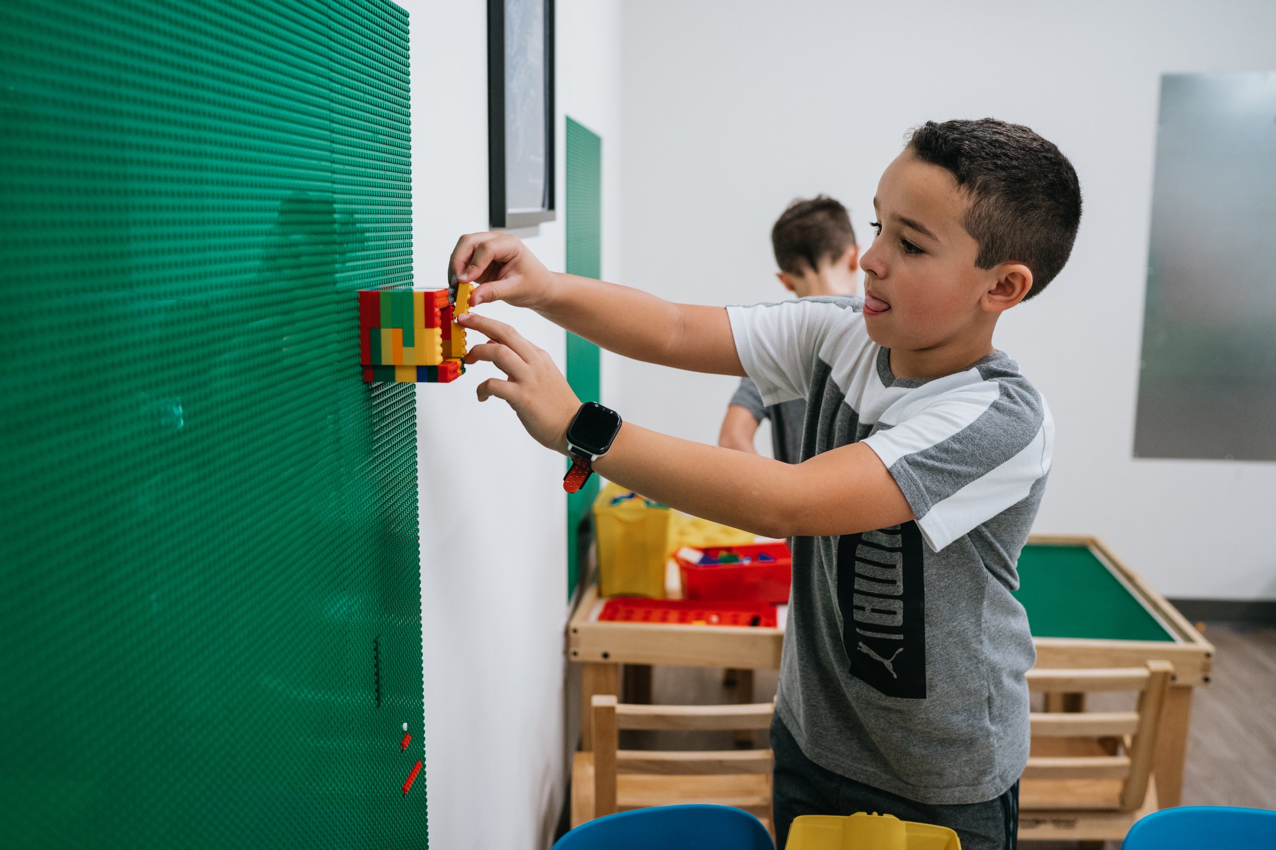 A young boy with short dark hair and a smartwatch on his left wrist is placing a colorful building block onto a green LEGO wall. In the background, another boy is visible, and there are furniture pieces including a yellow container, a red container, and a wooden table.