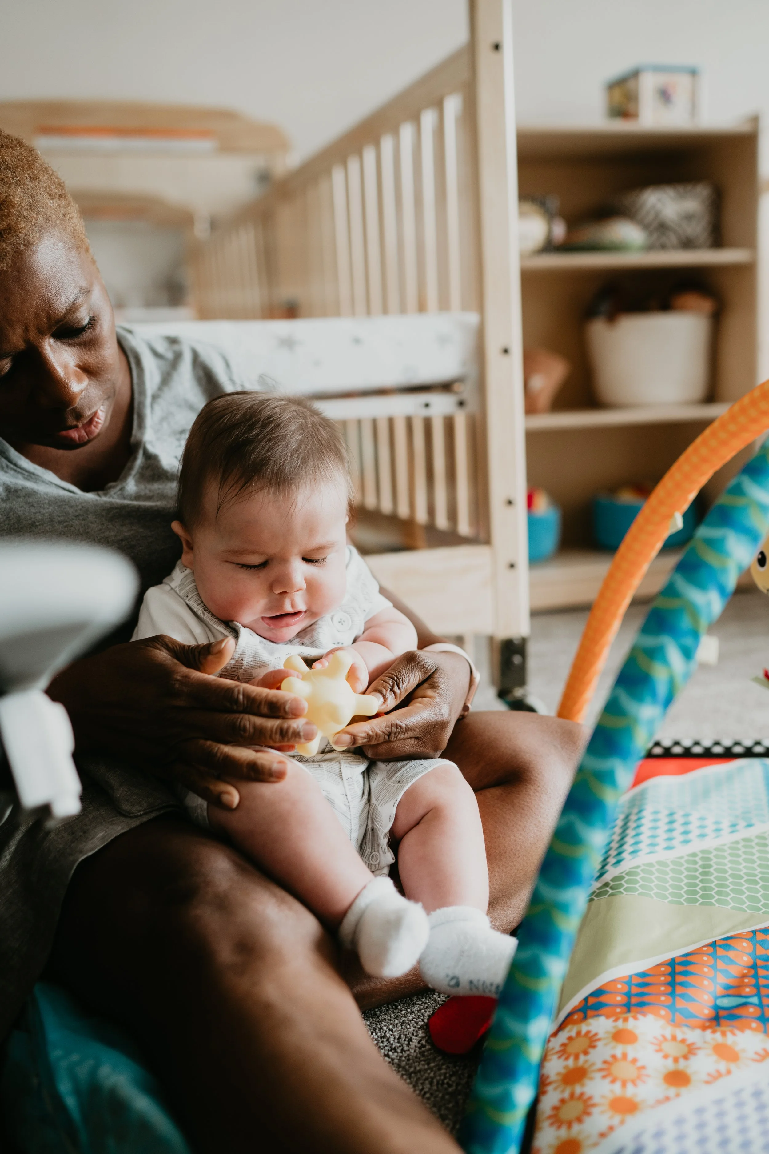 A woman holding a baby on her lap, playing with a toy in a nursery with a crib and shelves in the background.