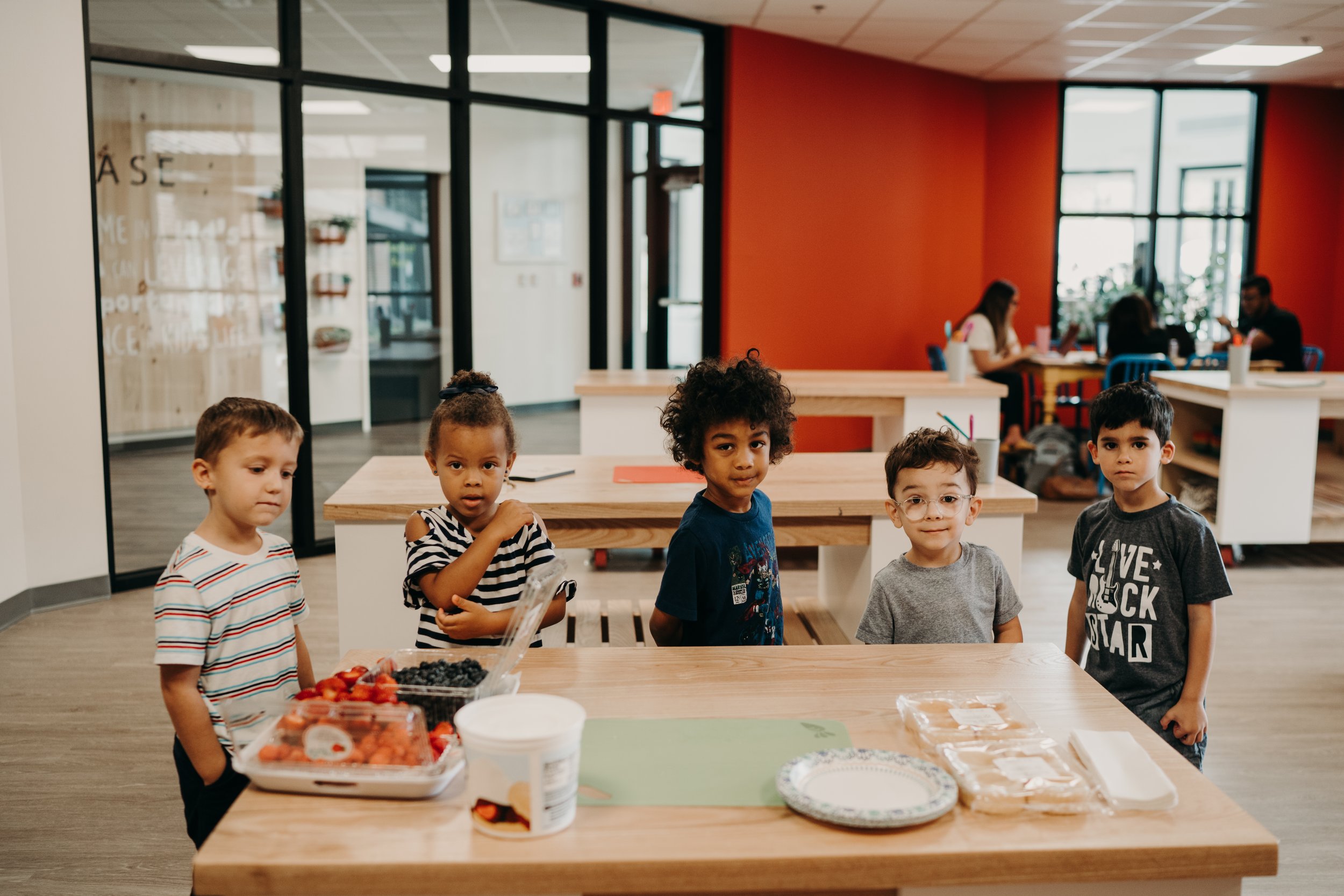 Five children standing behind a wooden table in a brightly lit room with other children and adults in the background, with plates of food and snacks on the table.