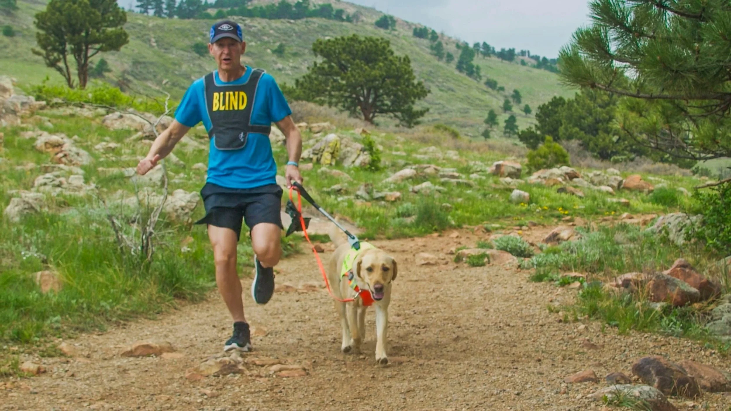 a man running with his seeing eye dog through the hills of boulder wearing a blind vest
