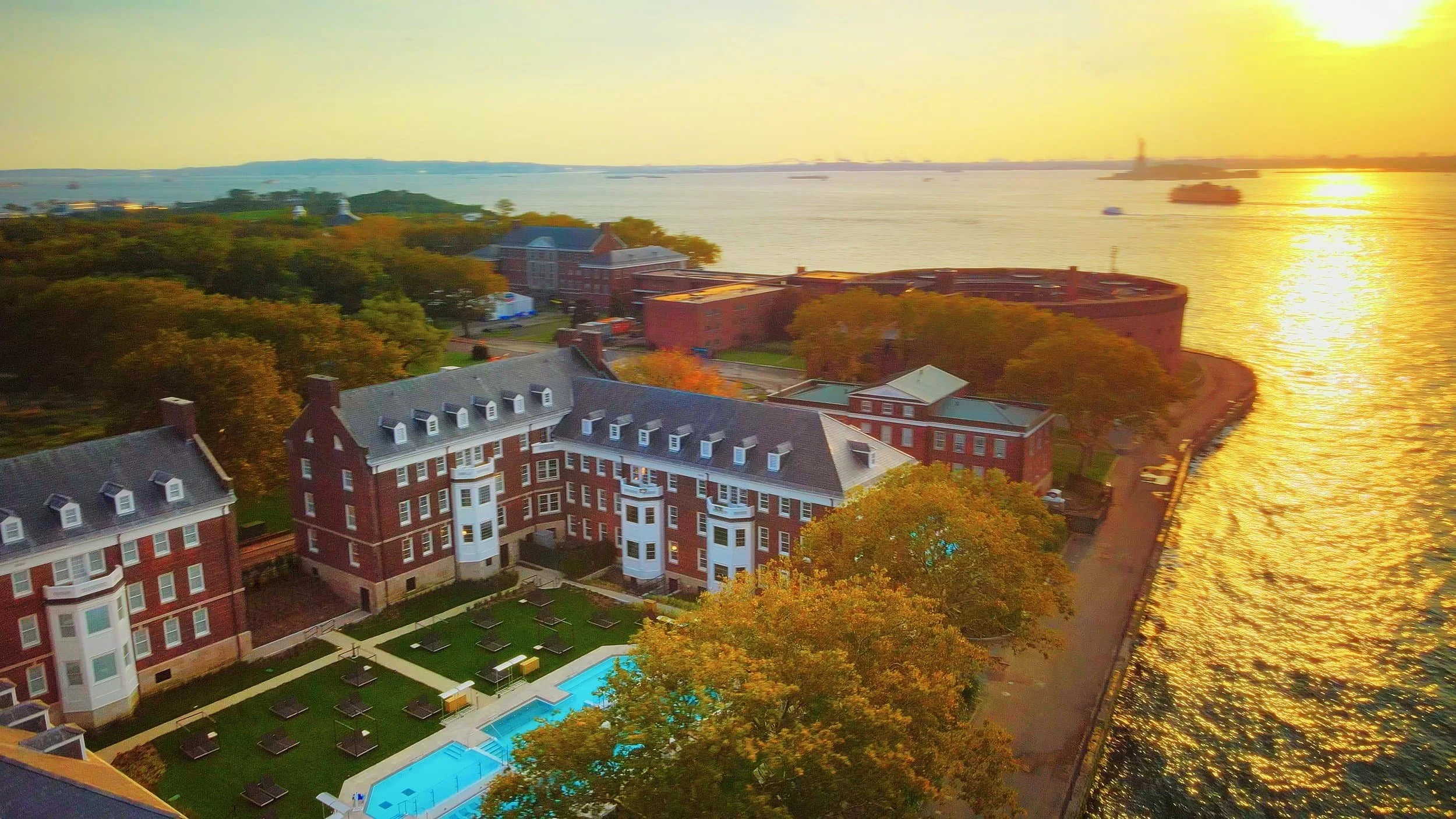 Aerial view of historic red brick buildings and a pool, overlooking a body of water at sunset with a golden sky.