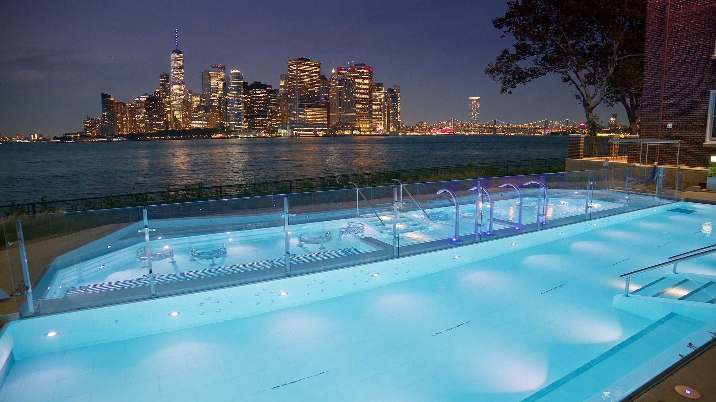 Night view of illuminated Manhattan skyline from a swimming pool with a glass barrier, overlooking a river and a bridge in the background.
