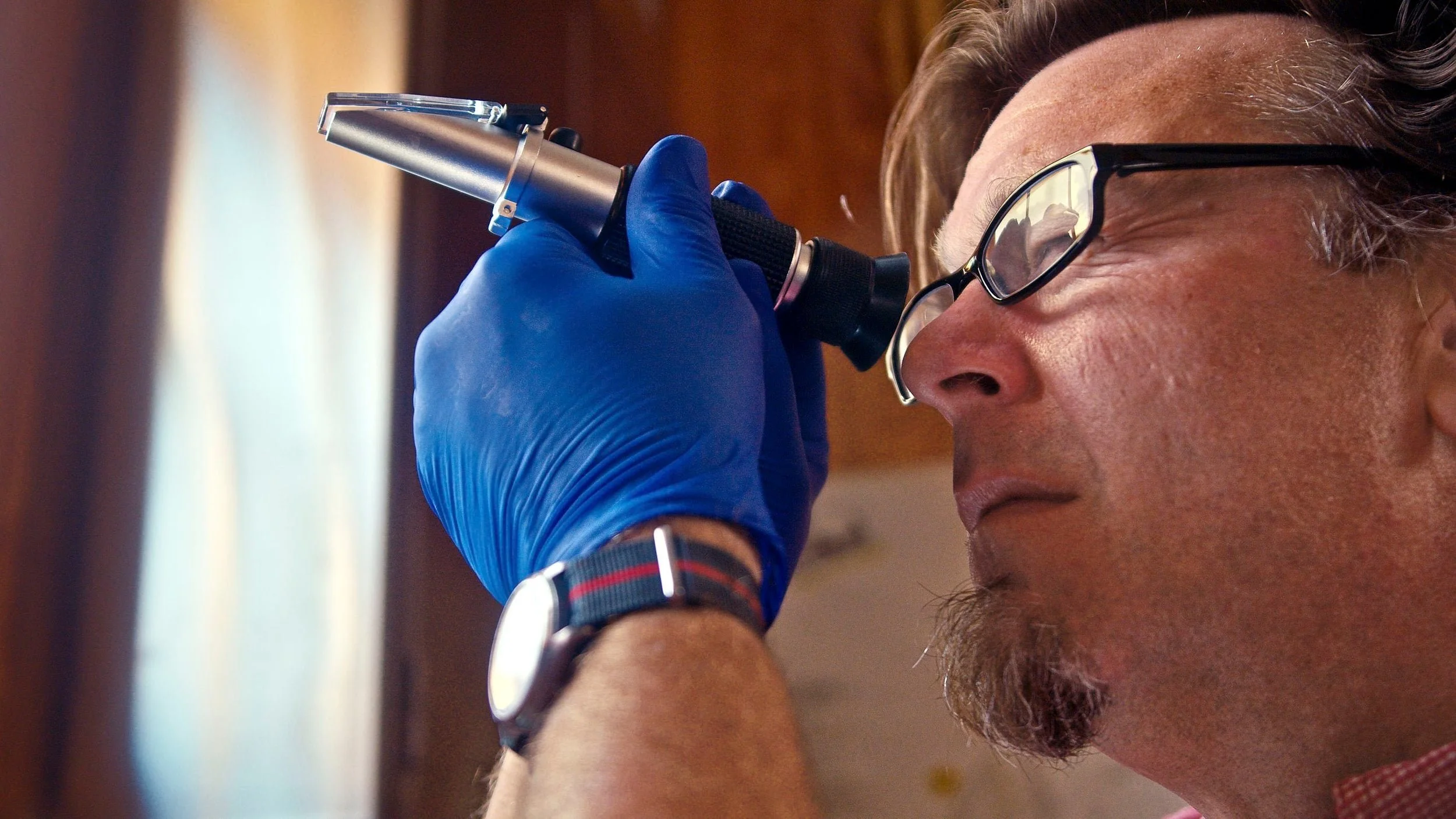 Close-up of a man with glasses and a beard, wearing a watch and a blue glove, looking into a microscope.