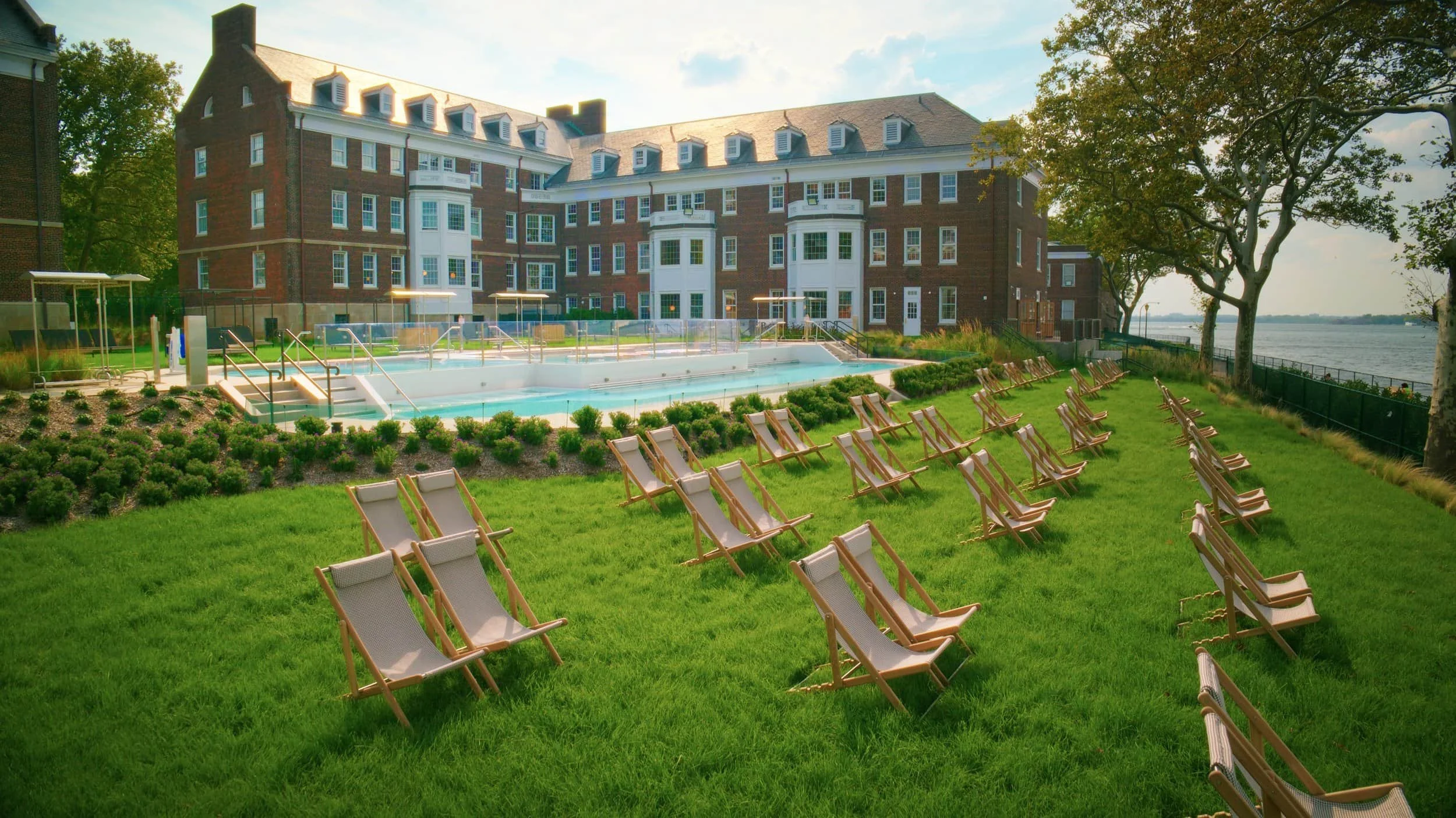 Lawn chairs arranged on a green grass lawn near a swimming pool at a large brick building with a view of a river, trees, and a partly cloudy sky.