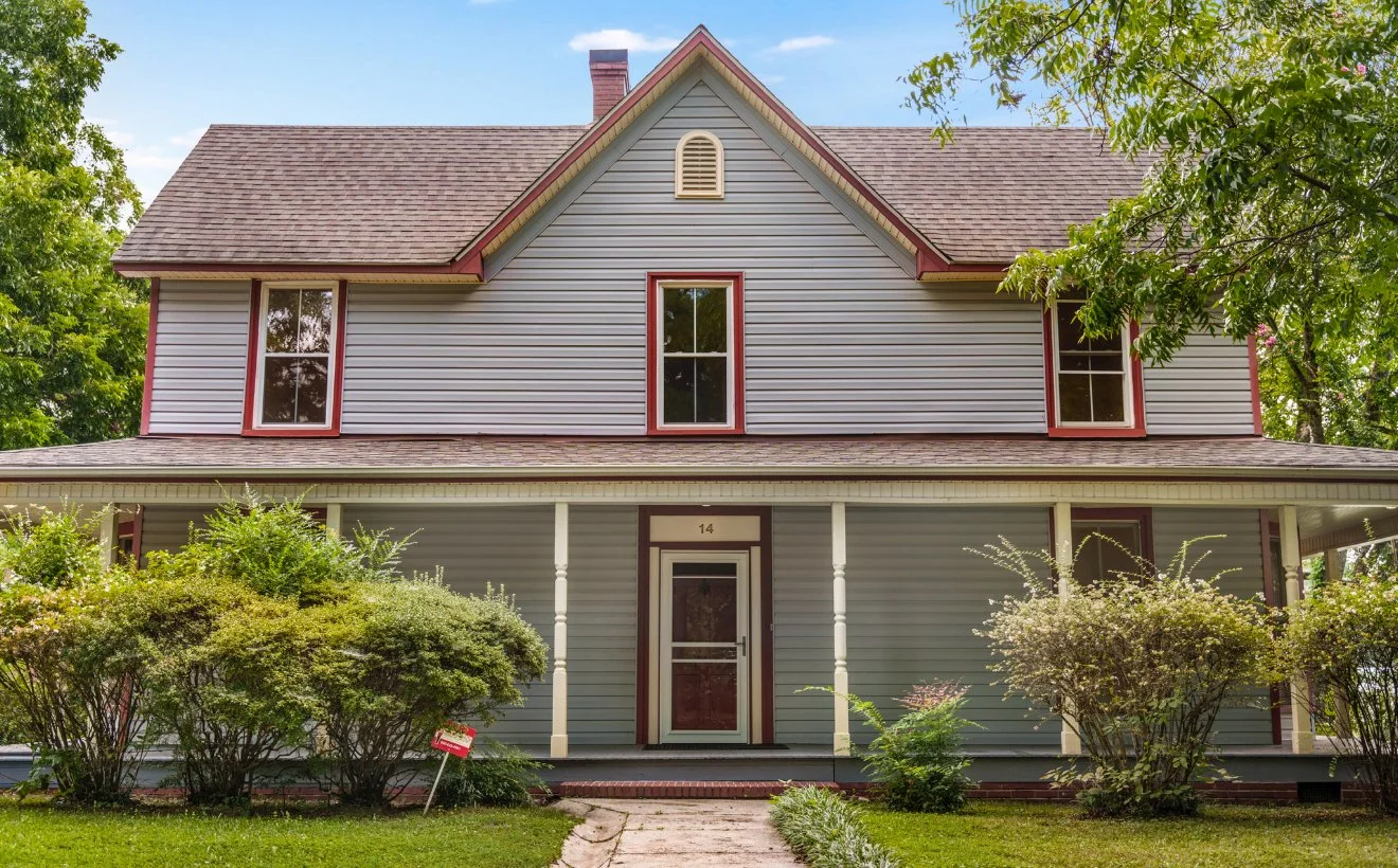 A two-story house with gray siding, red trim, a roof with shingles, and a front porch surrounded by bushes and trees.