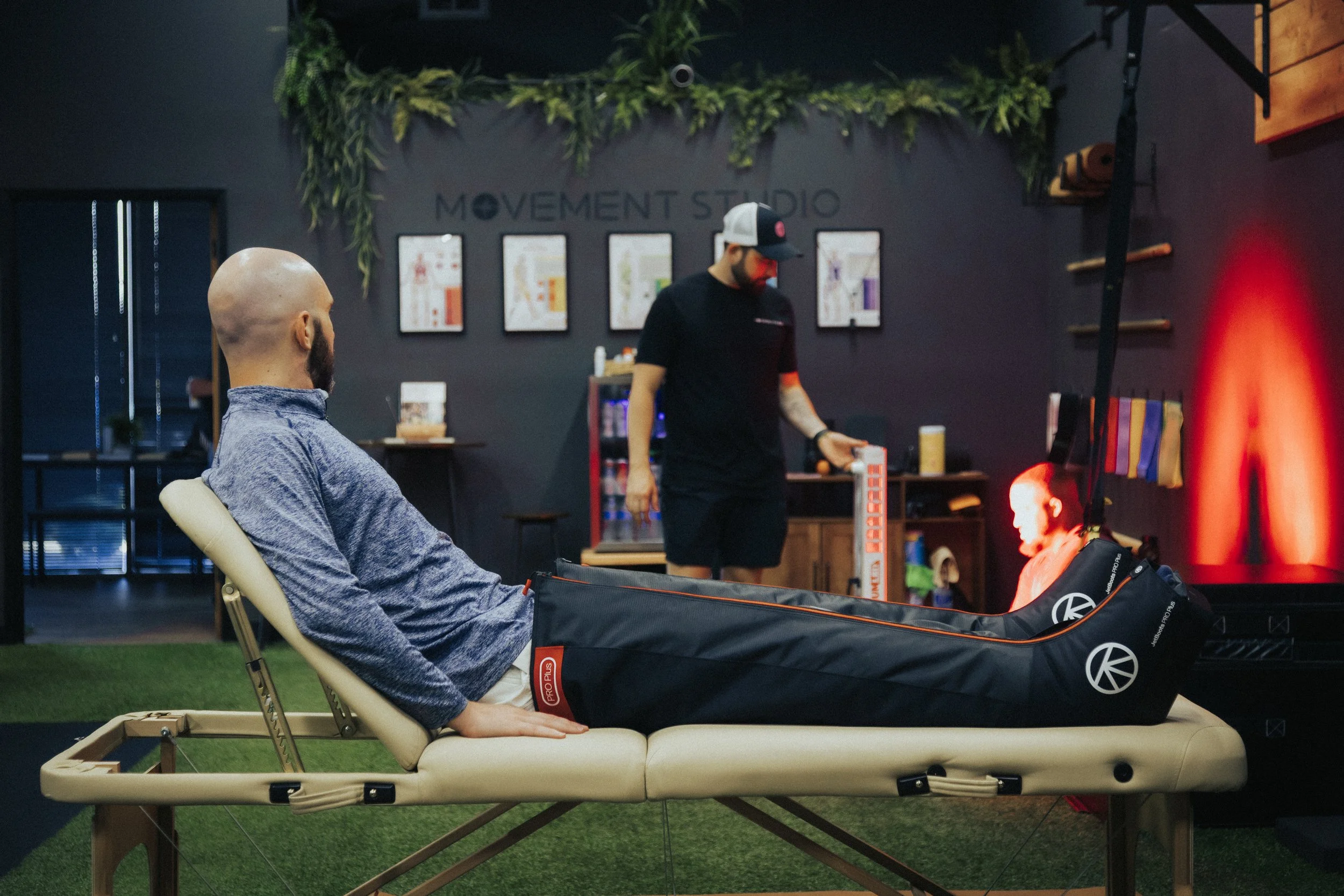 A man lying on a massage table with his legs elevated inside a fitness studio, with another man in black sportswear standing nearby, holding a foam roller and looking at a mannequin head with red LEDs. The background shows workout equipment and framed posters on a dark wall.