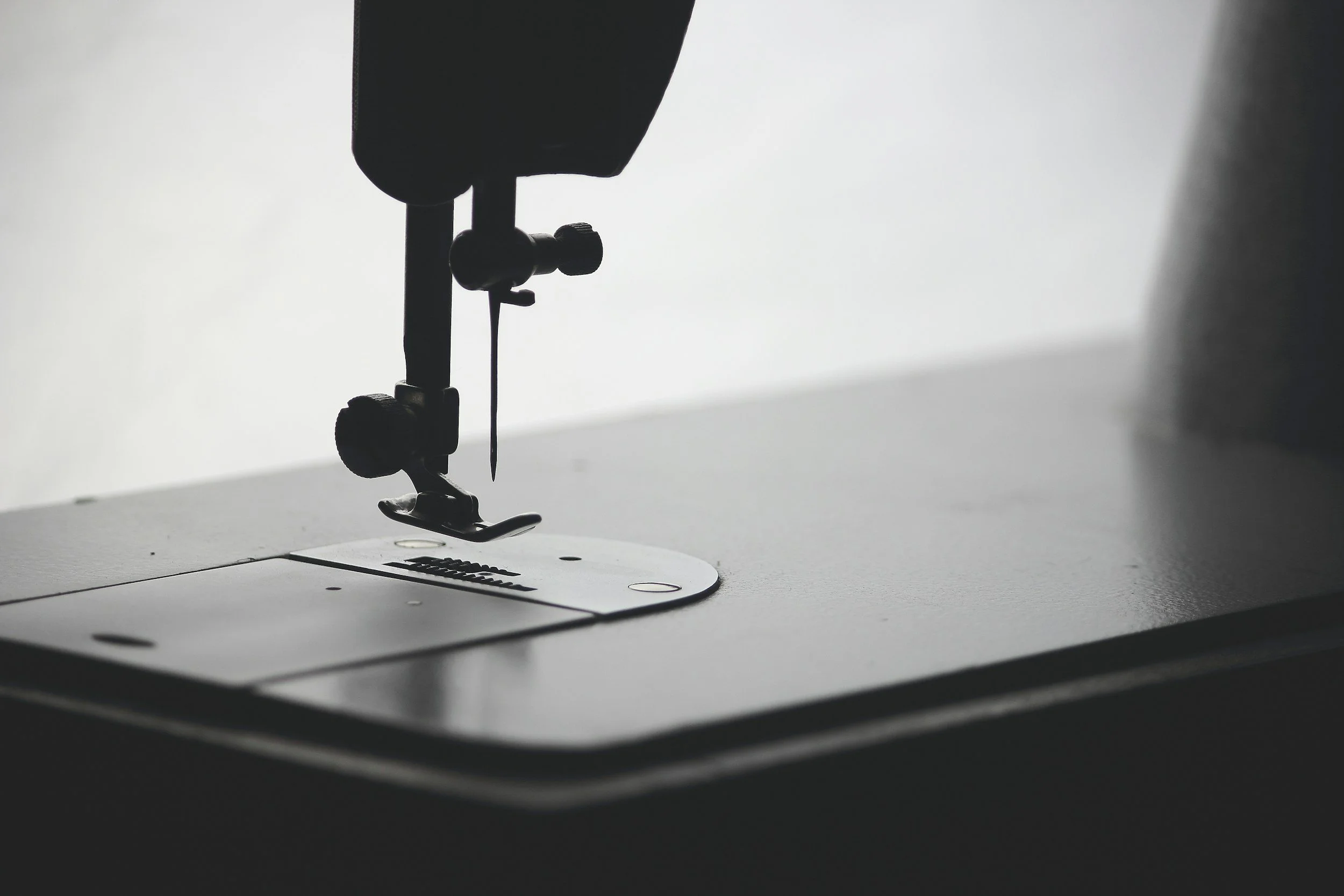 Close-up of a sewing machine needle and presser foot on a white sewing machine surface with a diffused light background.