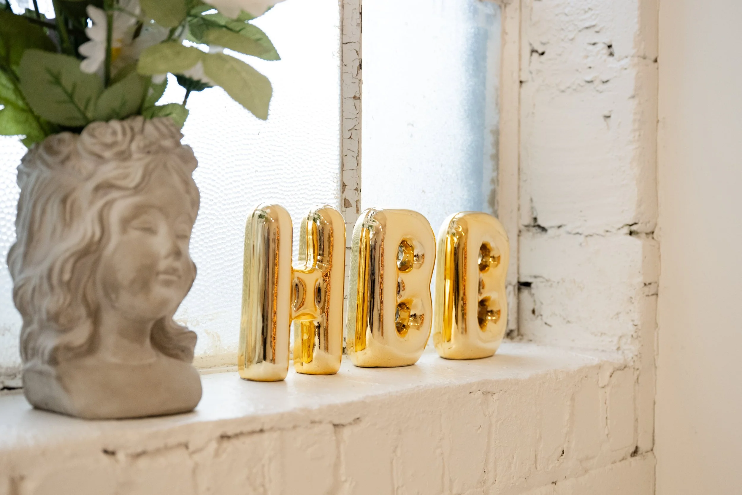 Decorative window sill with a beige bust sculpture of a woman's head, artificial green leaves in a flowerpot, and four gold-colored LED letter lights spelling 'LOVE' in a rustic white brick wall setting.