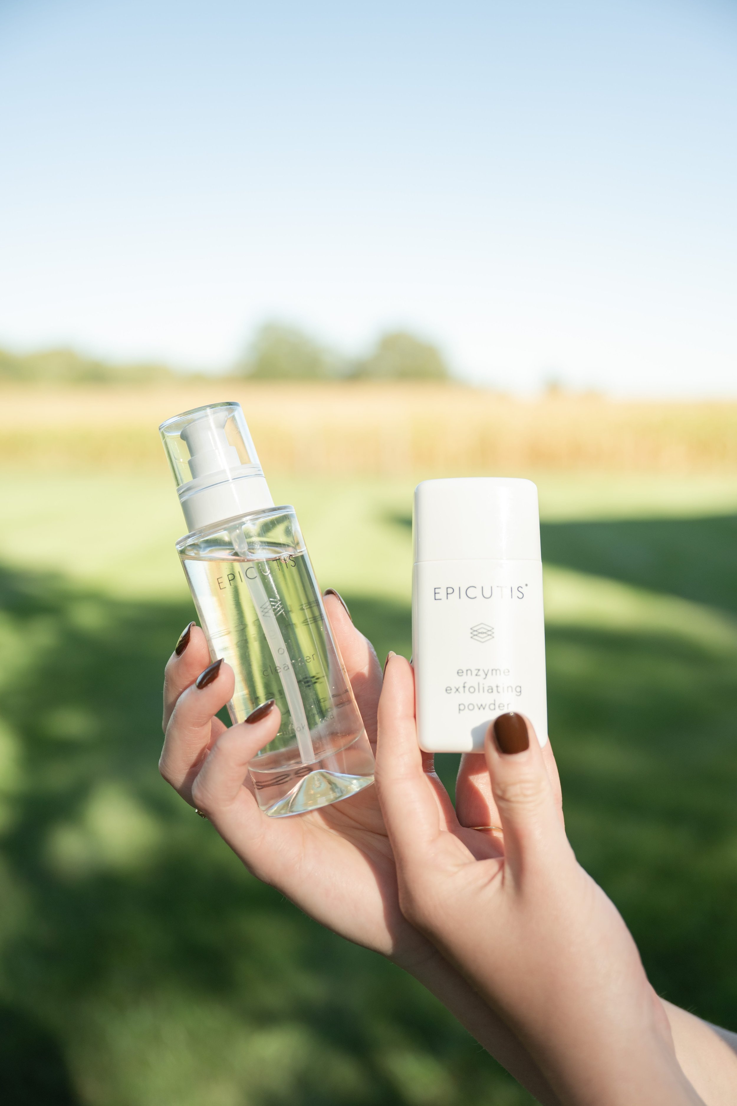 Hands holding a clear bottle of facial toner or essence and a white container of enzyme exfoliating powder outdoors on a sunny day.