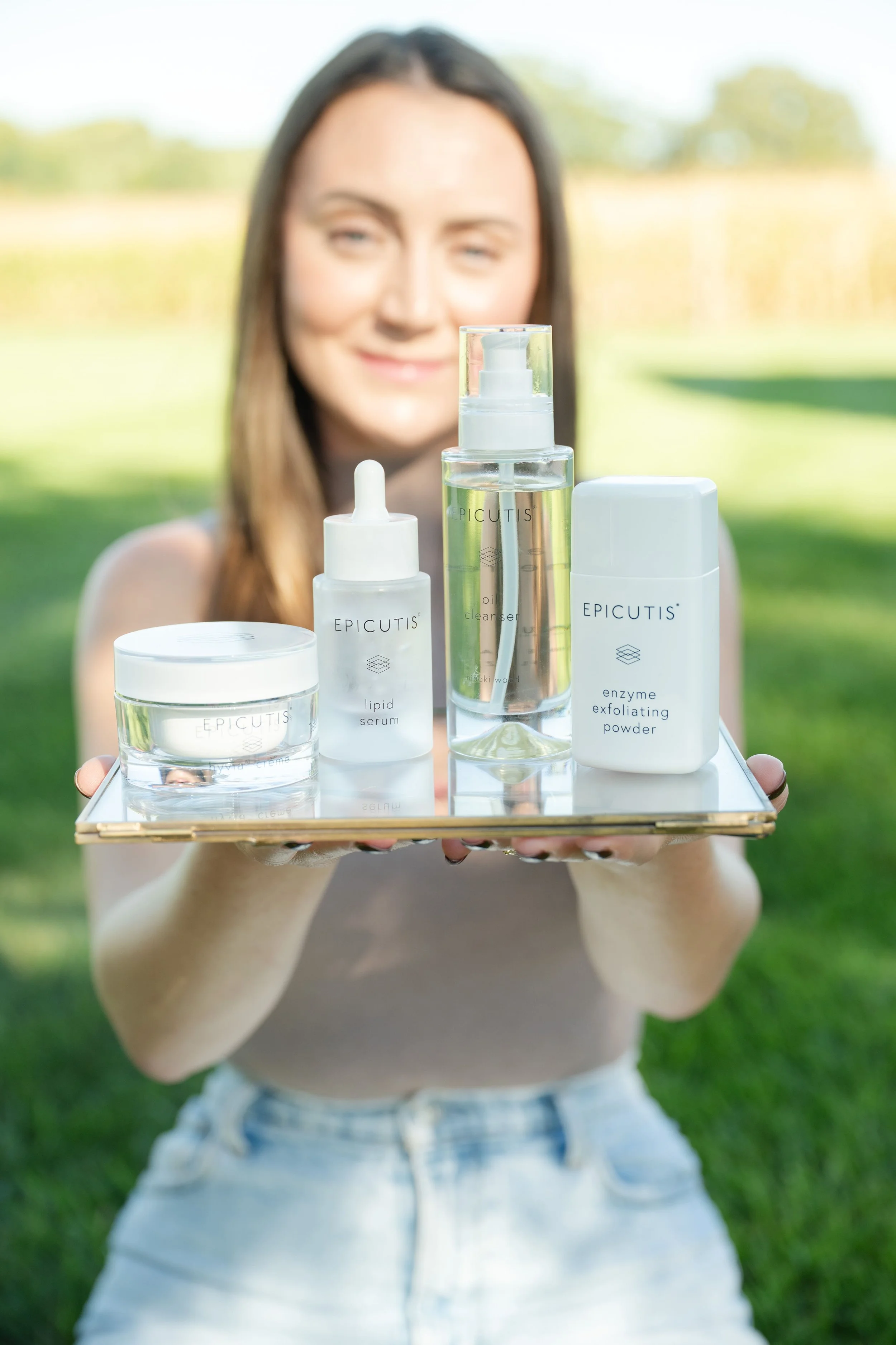 A woman holding a tray with skincare products outdoors, including a jar, dropper bottle, spray bottle, and exfoliating powder from the brand Epicuti's.