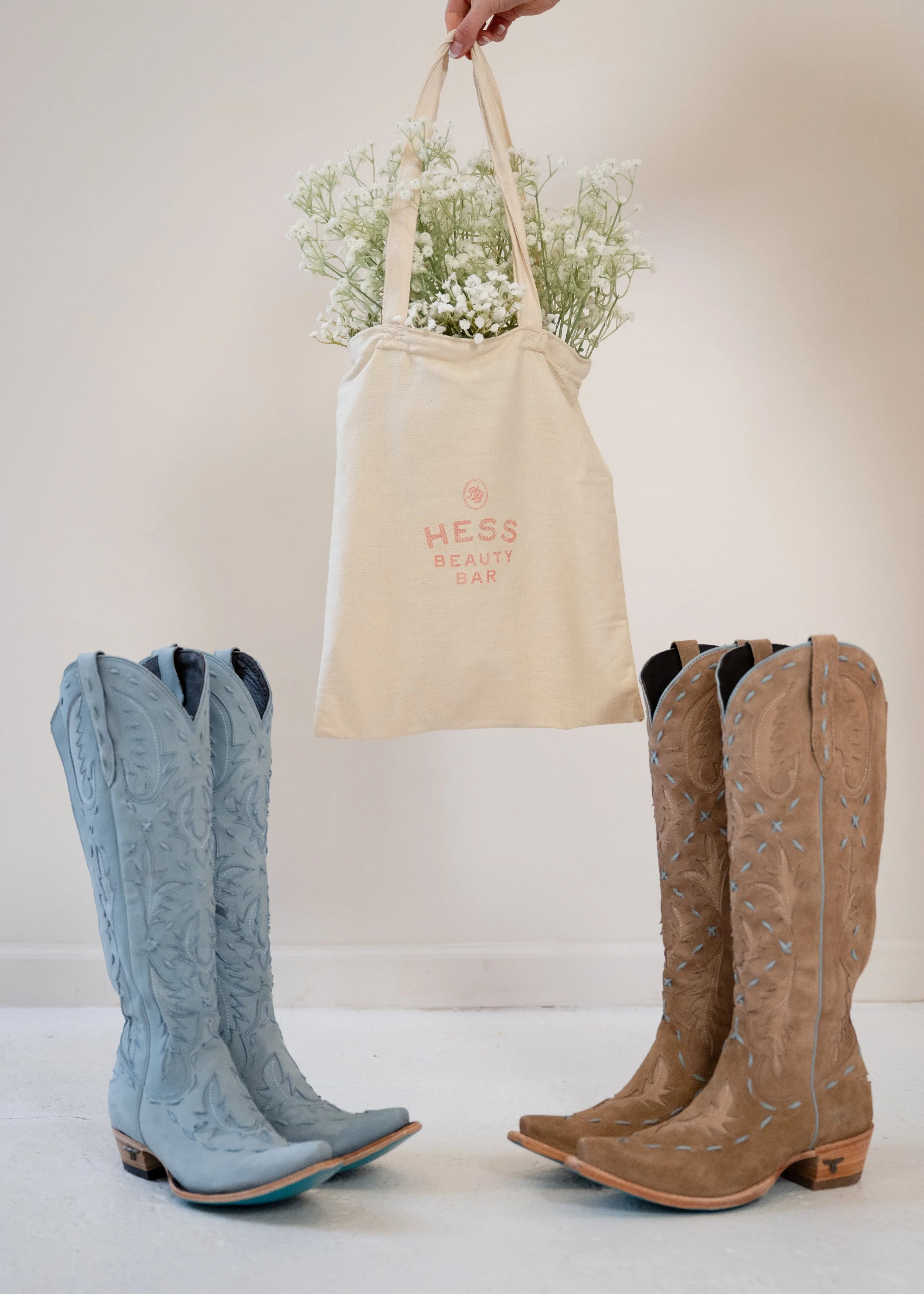 A person holding a beige tote bag with white flowers inside. The tote bag has red text that says 'HESS BEAUTY BAR'. There are two pairs of cowboy boots on the floor, one pair is light blue and the other is tan, both with decorative stitching.