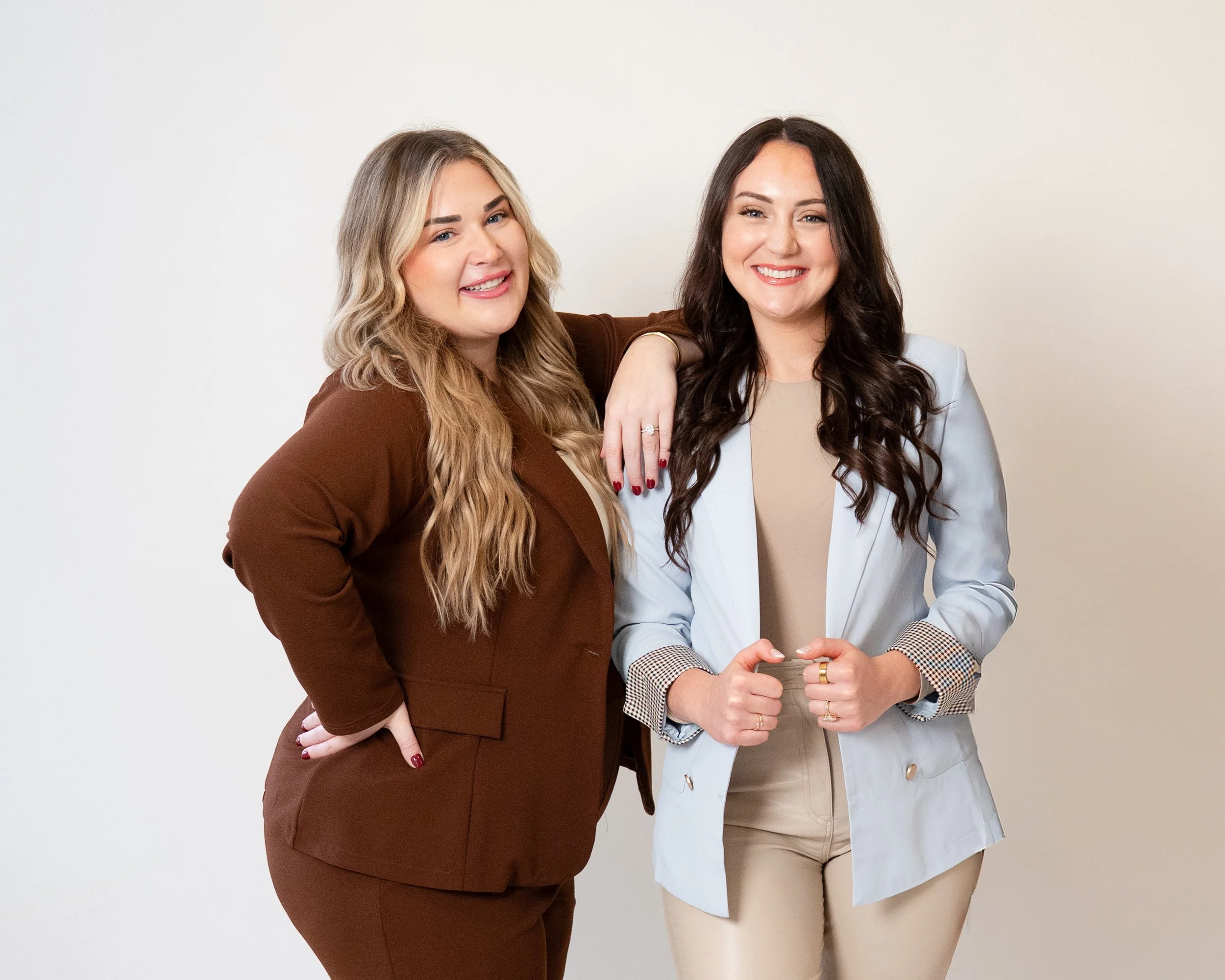 Two women smiling, one with blonde hair and the other with dark hair, standing against a plain background, with one woman resting her arm on the other's shoulder.