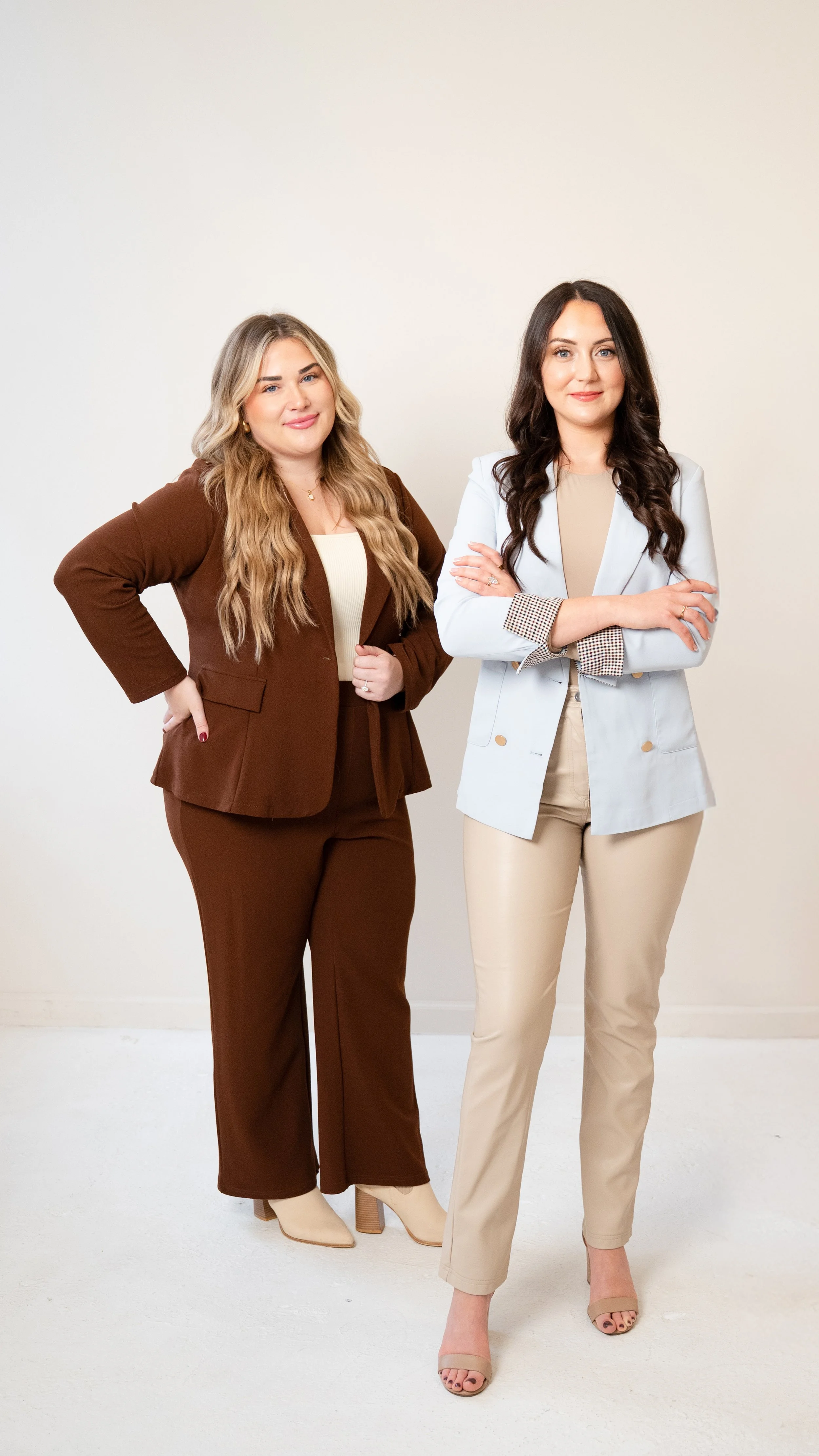 Two women standing against a plain wall, dressed in business casual attire, with confident expressions, one with crossed arms and the other with hand on her hip.