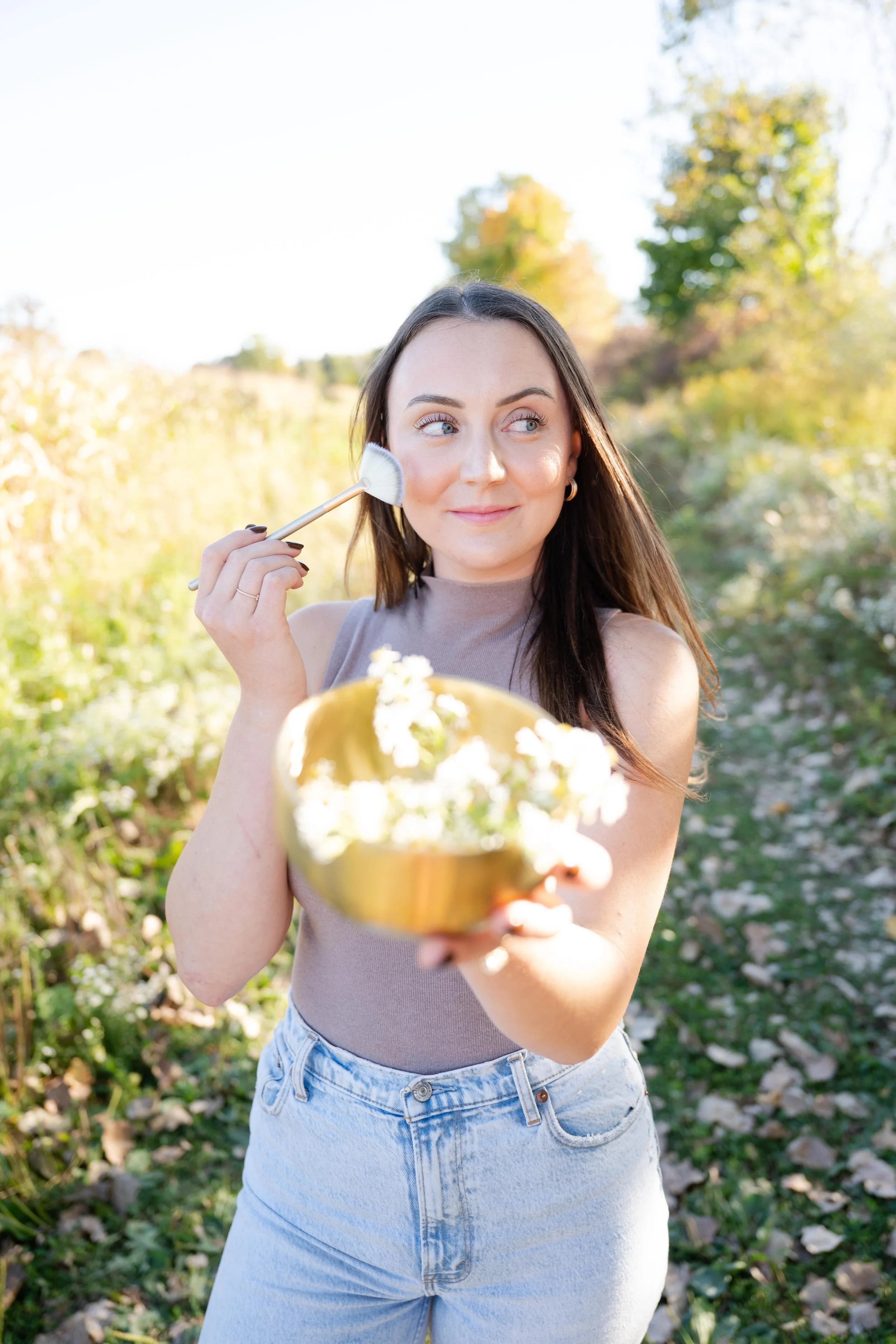 Young woman outdoors holding a gold bowl with white flowers, applying powder to her face with a makeup brush, surrounded by autumn foliage.