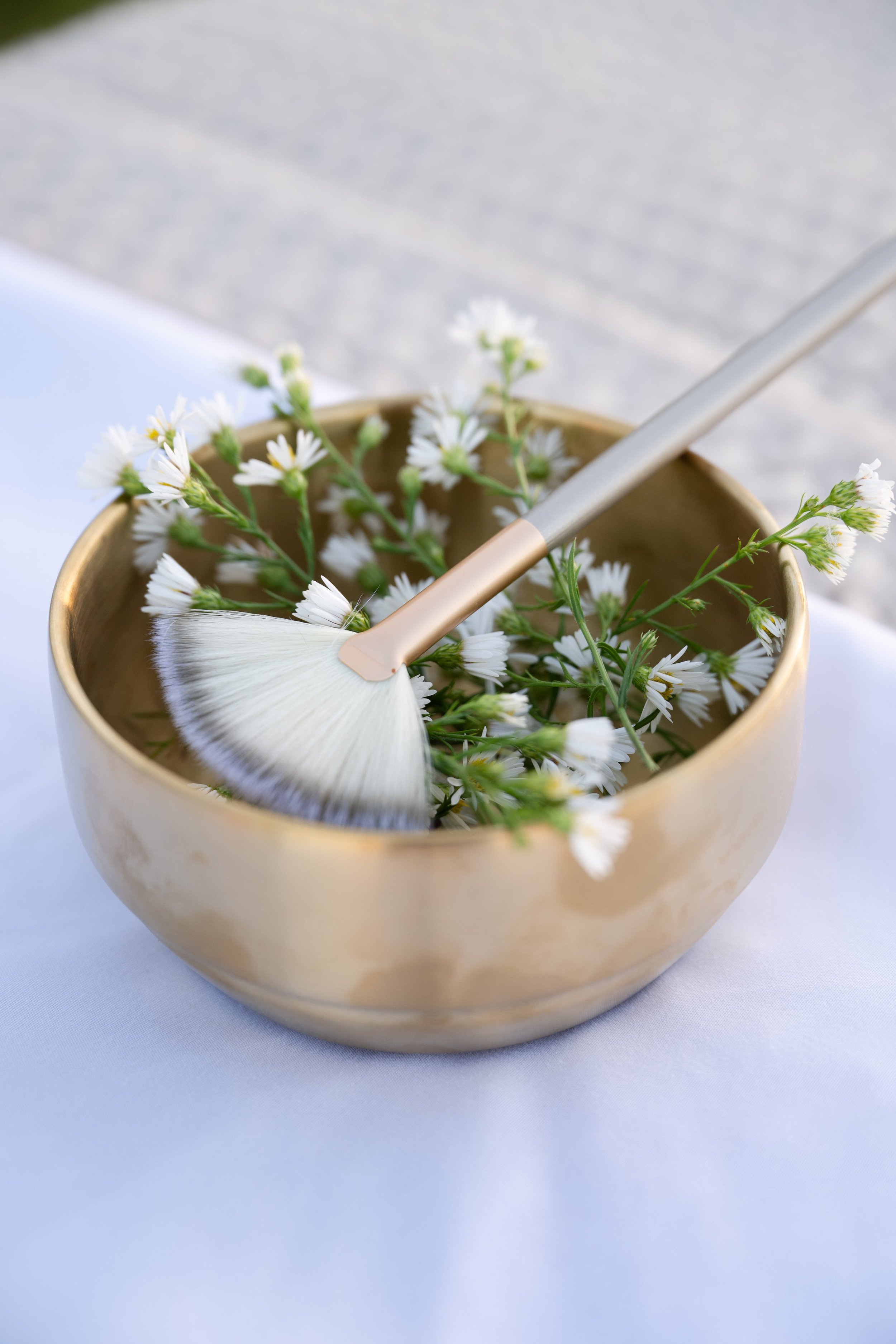 A gold bowl filled with small white flowers and a brush for cleaning, placed on a white cloth surface.
