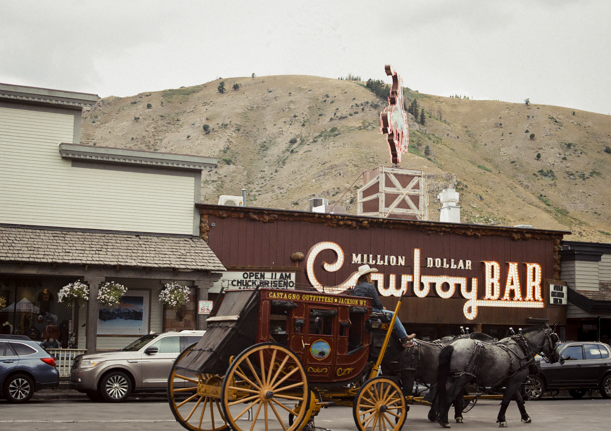 A horse-drawn carriage in front of a bar with a neon sign reading 'Million Dollar Cowboy Bar'. There are cars parked and a mountain in the background.