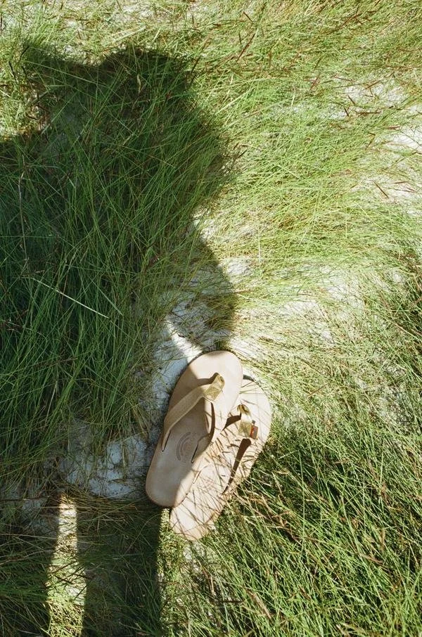 A pair of tan sandals lying on sandy ground amid tall green grass, with a person's shadow cast nearby.