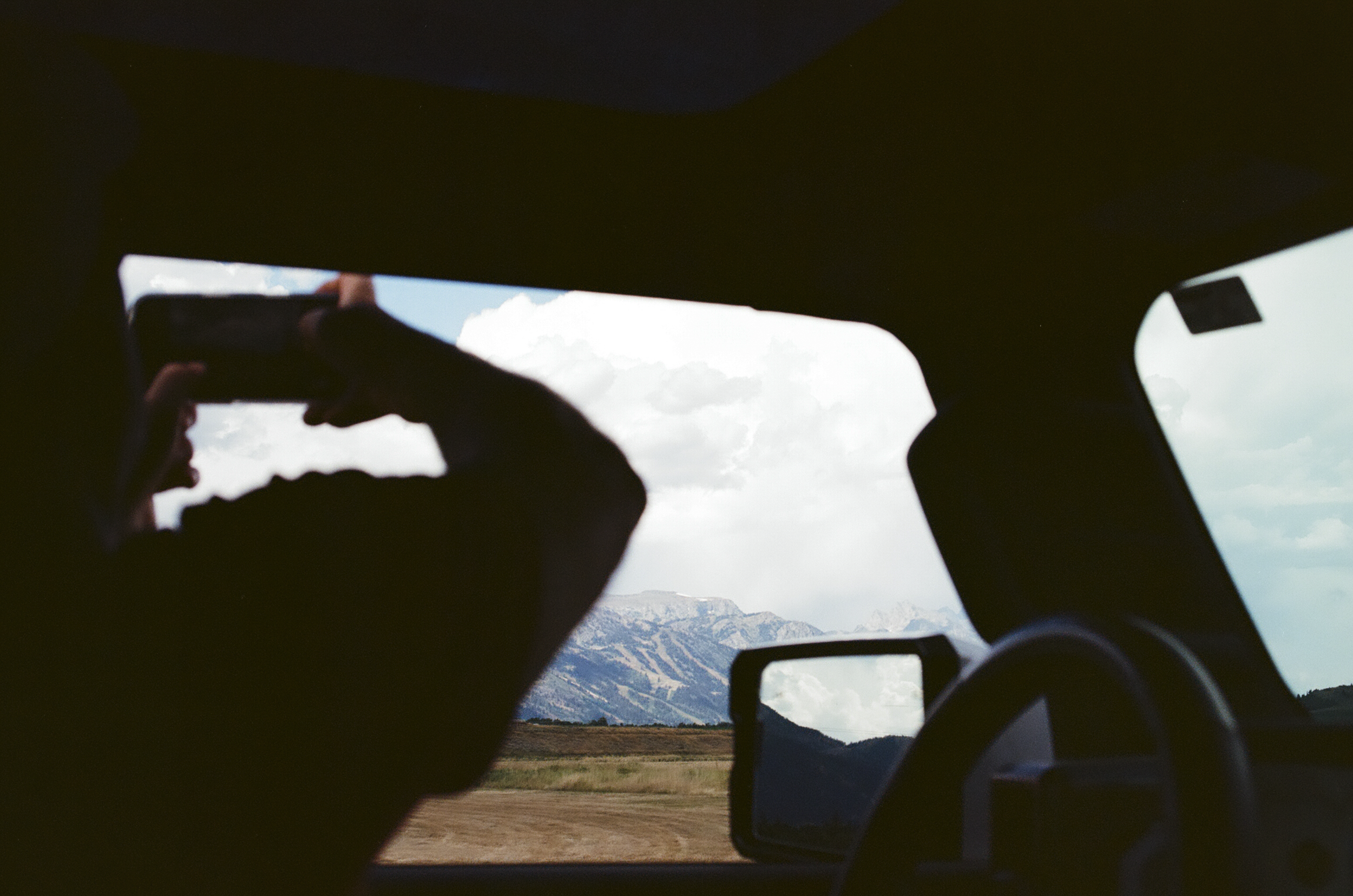 Person taking a photo outside the vehicle through the rear window with a camera or phone, scenic landscape with mountains and clouds in the background.
