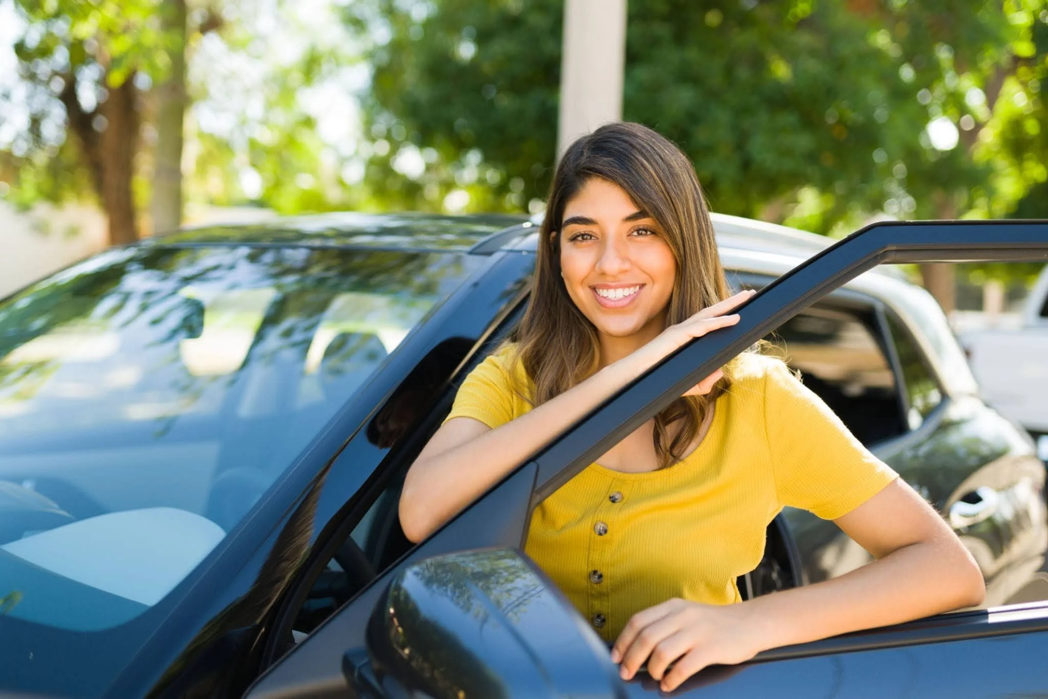 A young woman in a yellow shirt smiling and leaning out of a car window, parked outdoors with trees in the background.