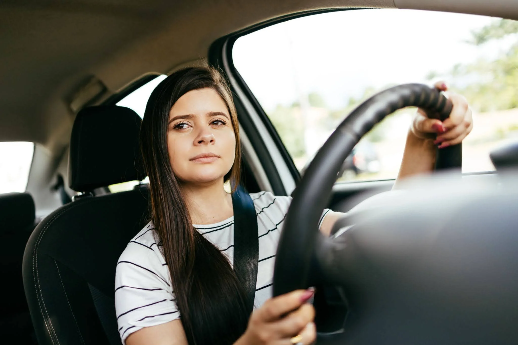 Young woman with long dark hair driving a car, holding the steering wheel with both hands, wearing a striped T-shirt, looking confident, with a seatbelt on, outside scenery visible through the window.