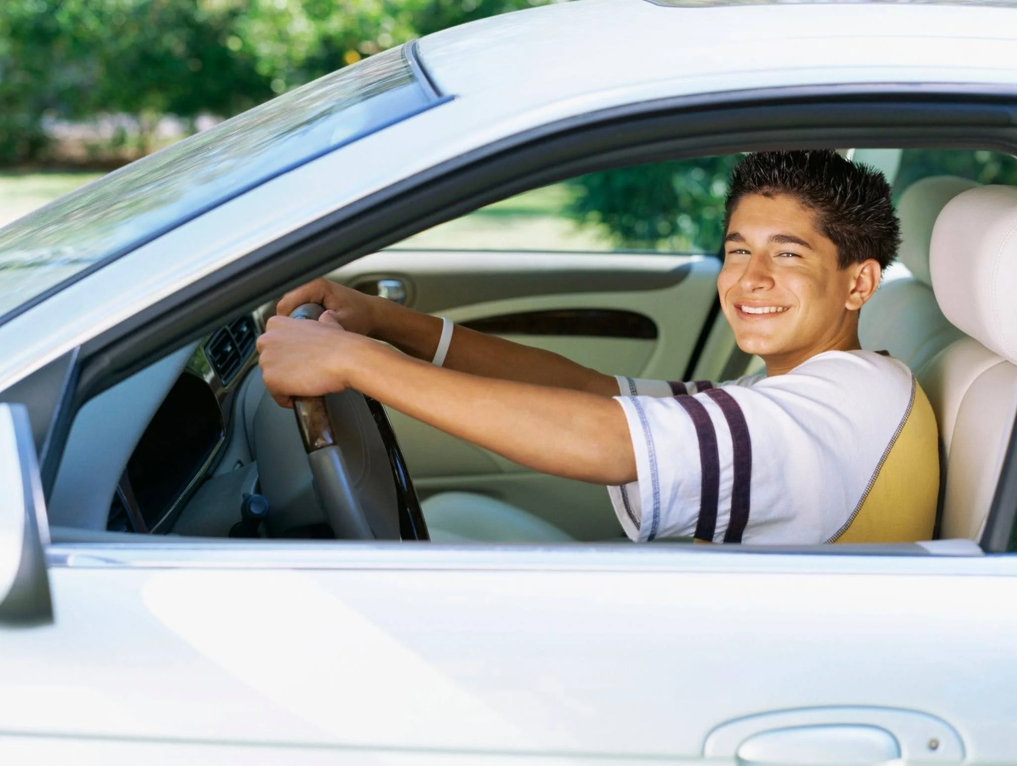 Young man smiling while driving a white car on a sunny day in a green park.