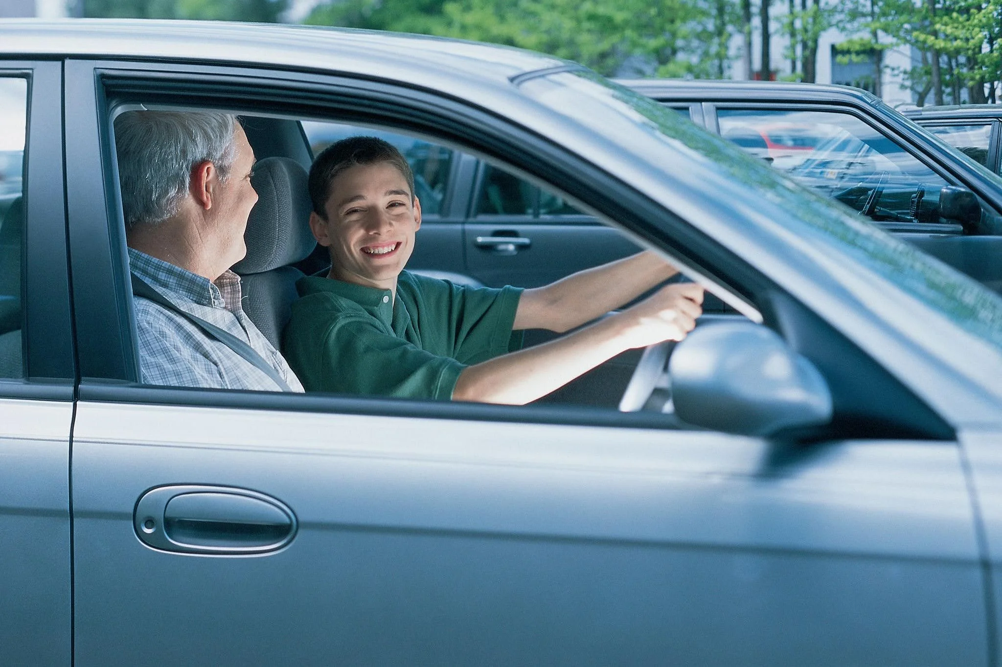 A young boy and an older man sitting in a car, smiling at each other. The boy is in the driver’s seat, and the man is in the passenger seat. They appear to be having a conversation, with a parking lot and trees visible outside.